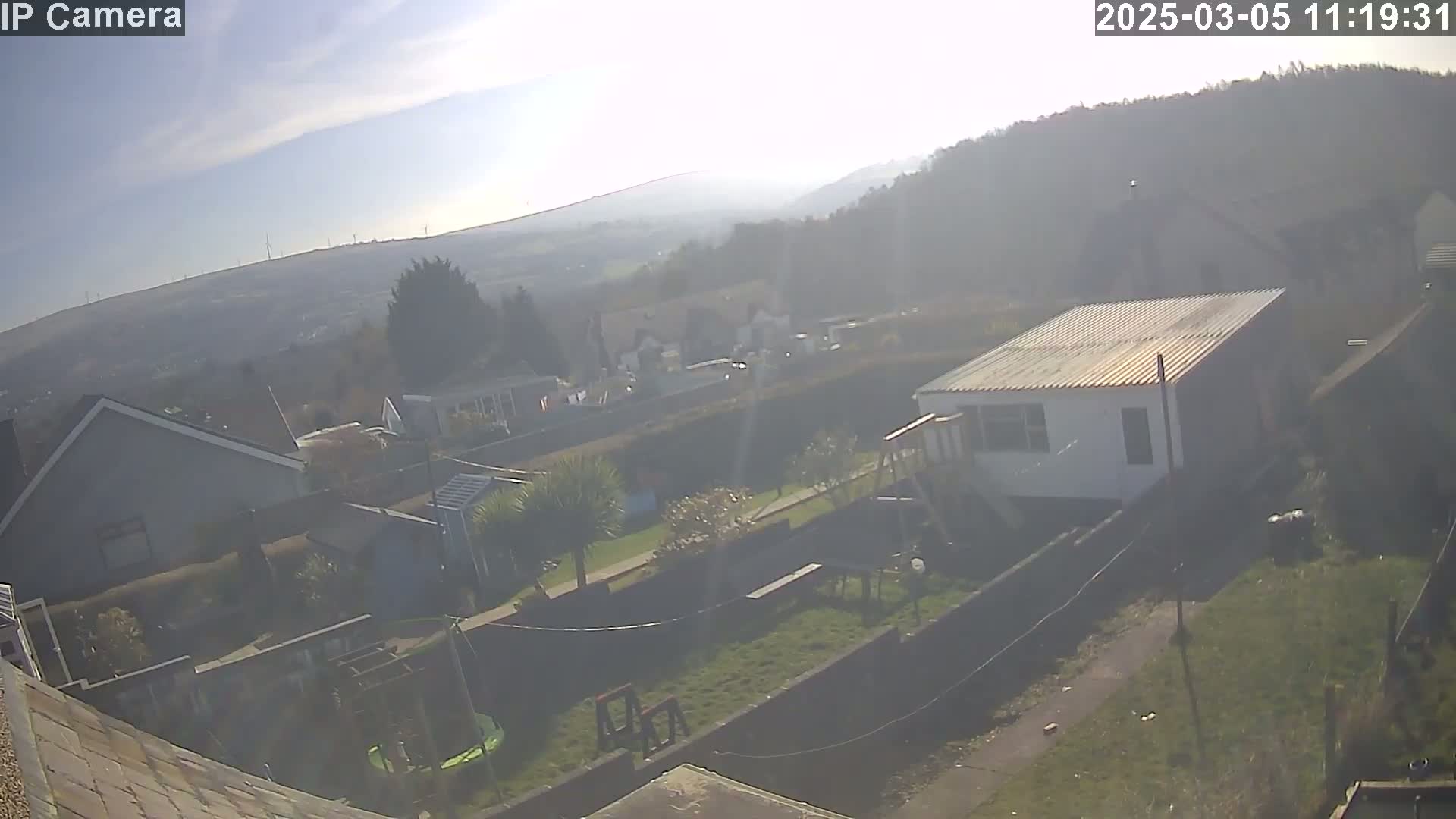 A hazy, sunny day overlooks a residential area with houses, gardens, and a play set, showing a view of distant hills and wind turbines.