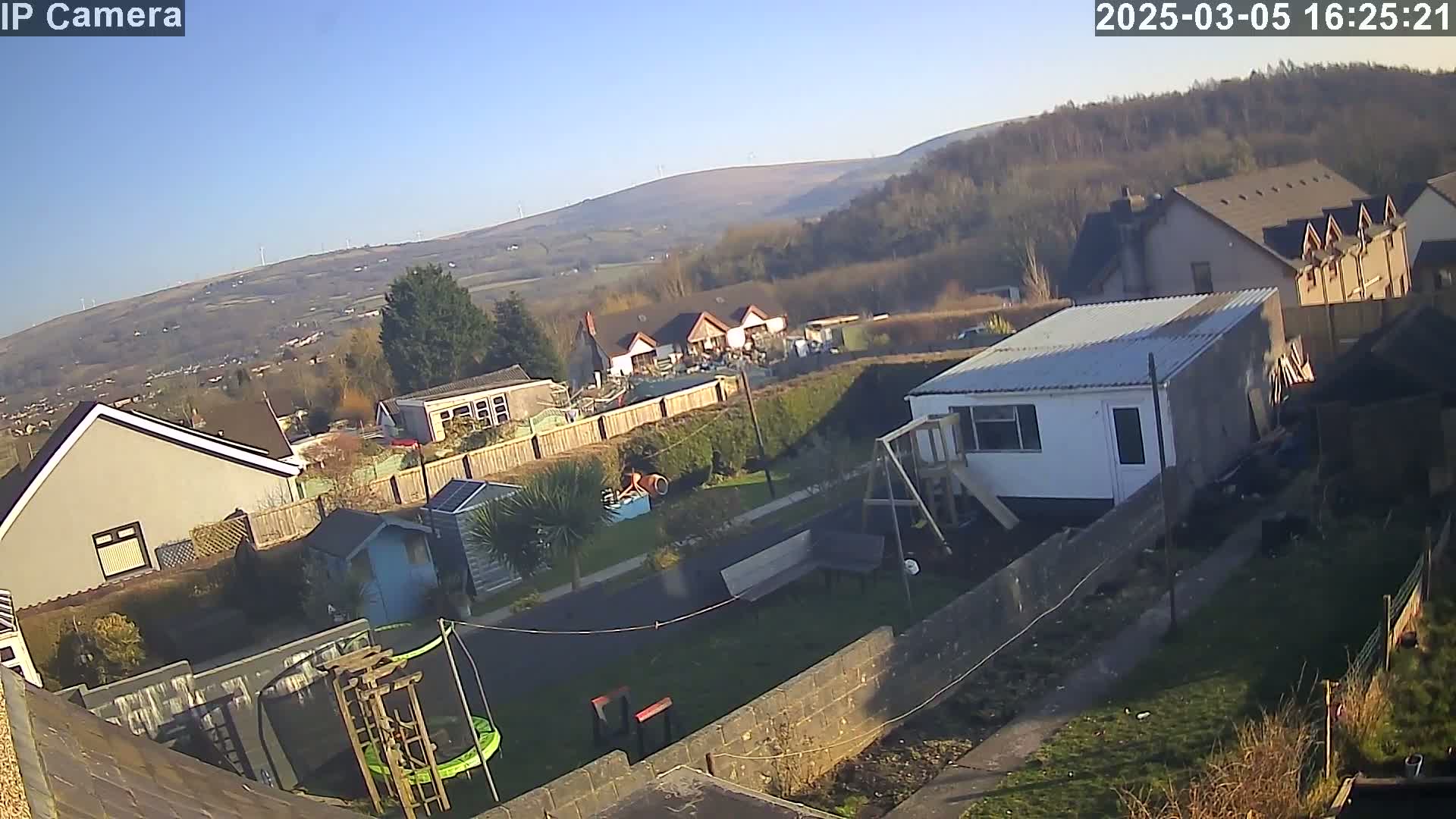 A backyard scene on a sunny day shows houses, a trampoline, a playset, and a small shed, with distant hills and wind turbines visible in the background.
