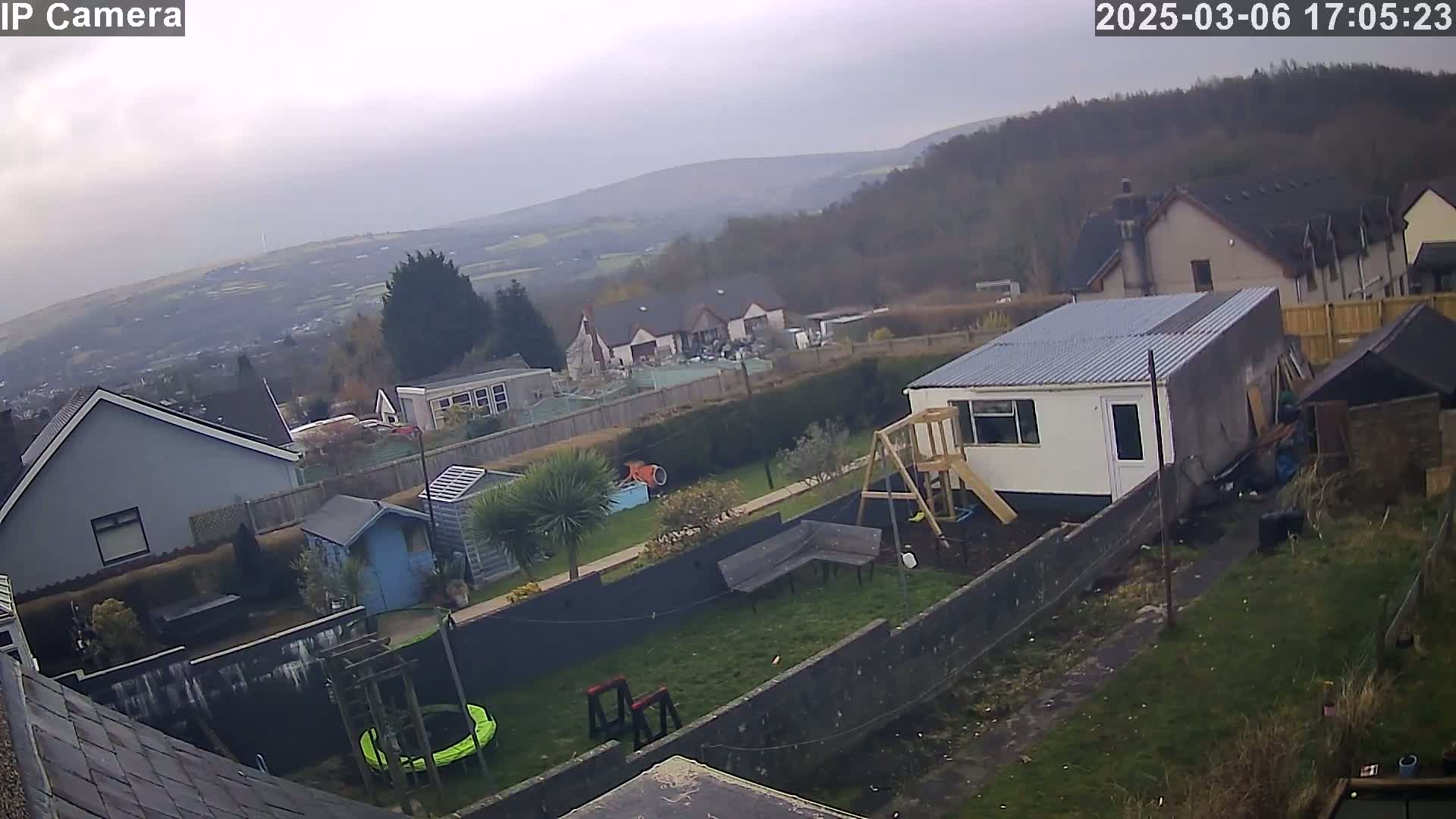 An overcast day reveals a backyard scene with a house, garden structures, a playset, and a trampoline, all set against a backdrop of distant hills and houses.