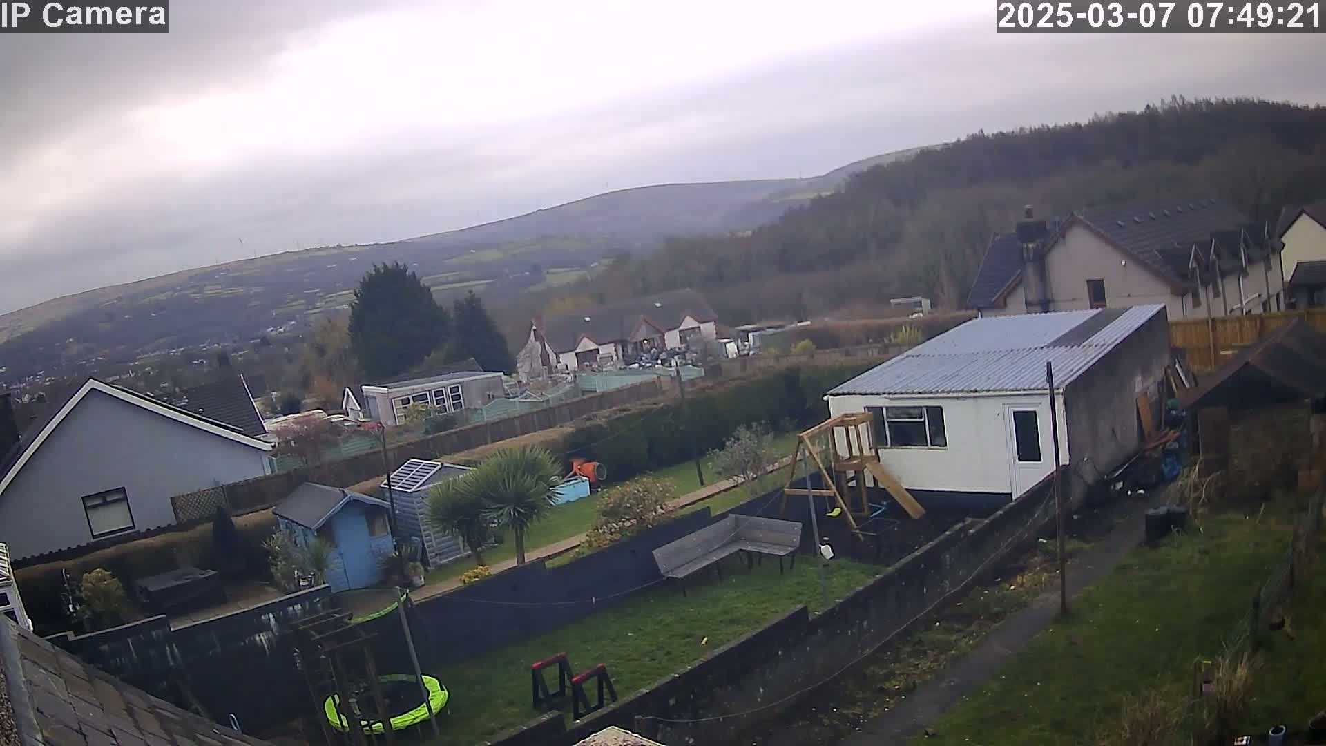 A backyard scene on an overcast day shows a garden with a shed, playset, and trampoline, overlooking houses and distant hills.