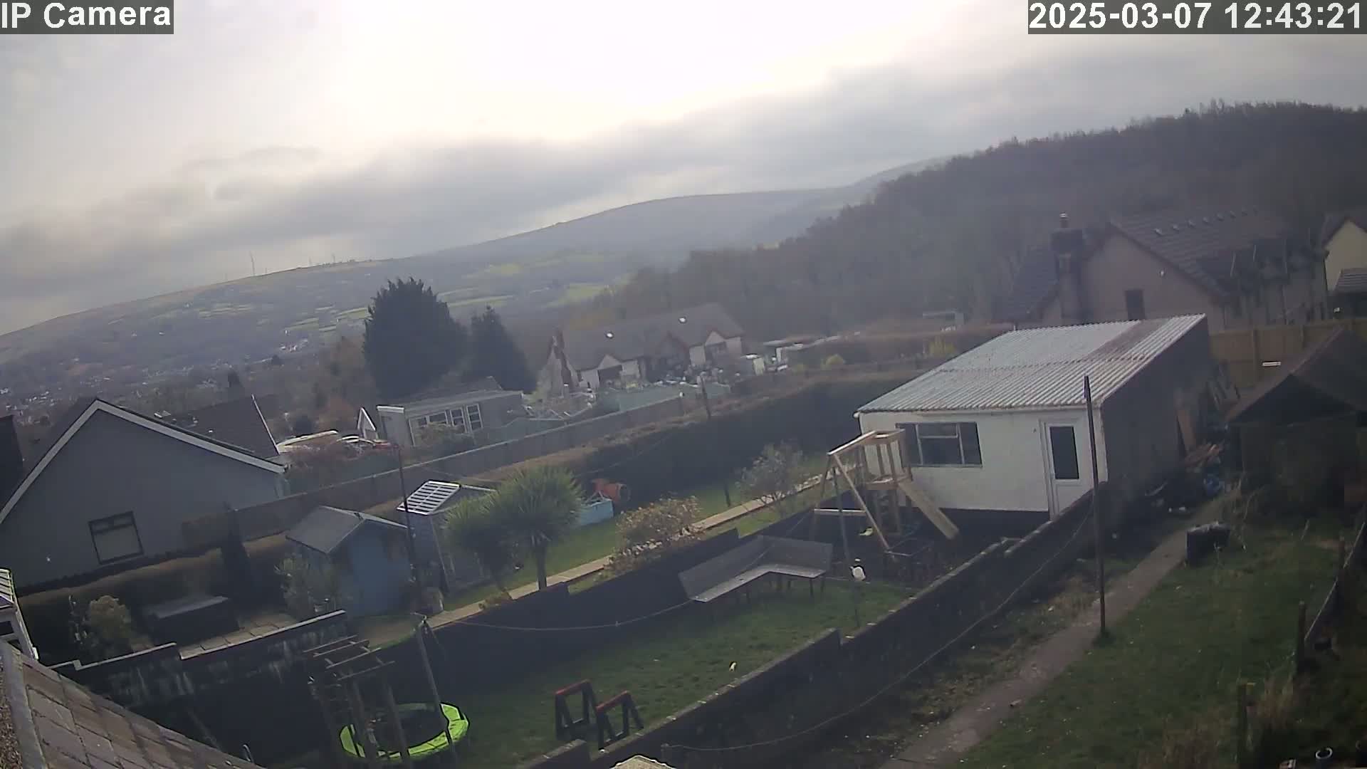 A cloudy day reveals a view of houses and gardens, with a hill and distant town visible in the background.