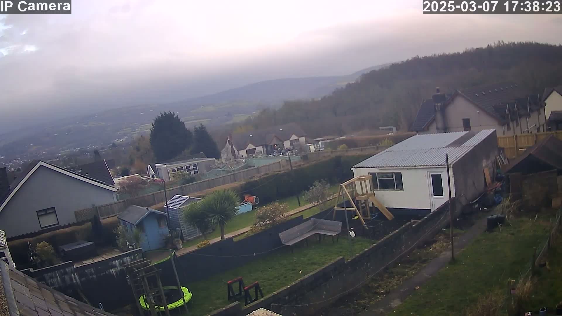 An overcast day reveals a backyard scene with houses, a garden, and a small shed, all overlooking a valley and distant hills.