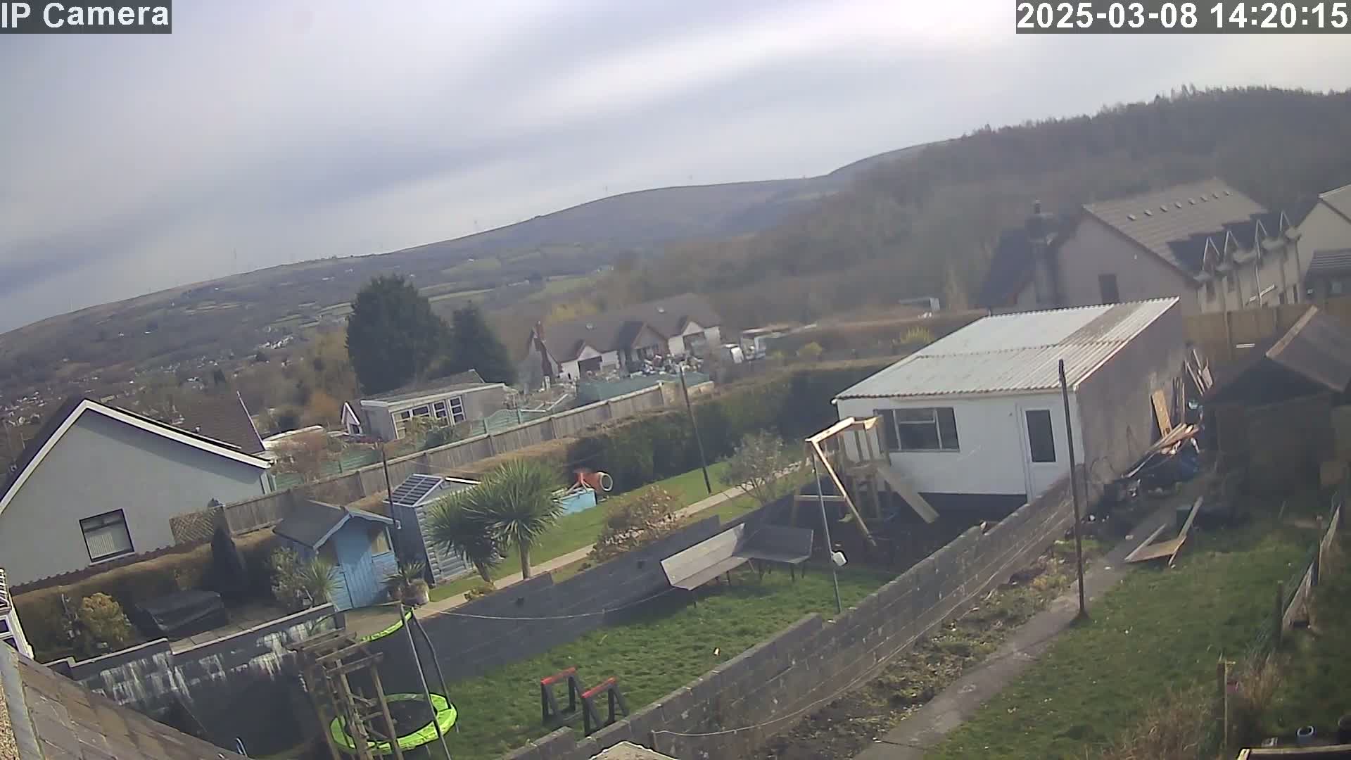A backyard with a trampoline, shed, and playset overlooks houses and a hilly landscape under a partly cloudy sky.