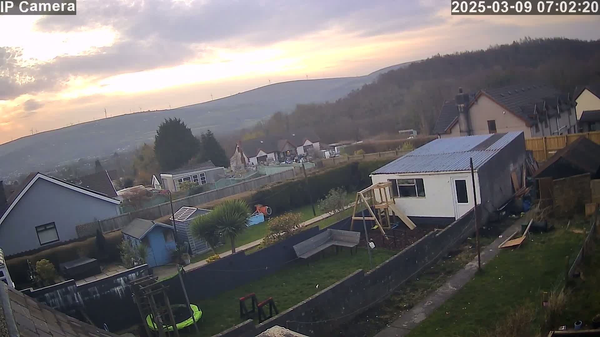 A backyard with houses and a hilly landscape in the background under a partly cloudy sunset sky.