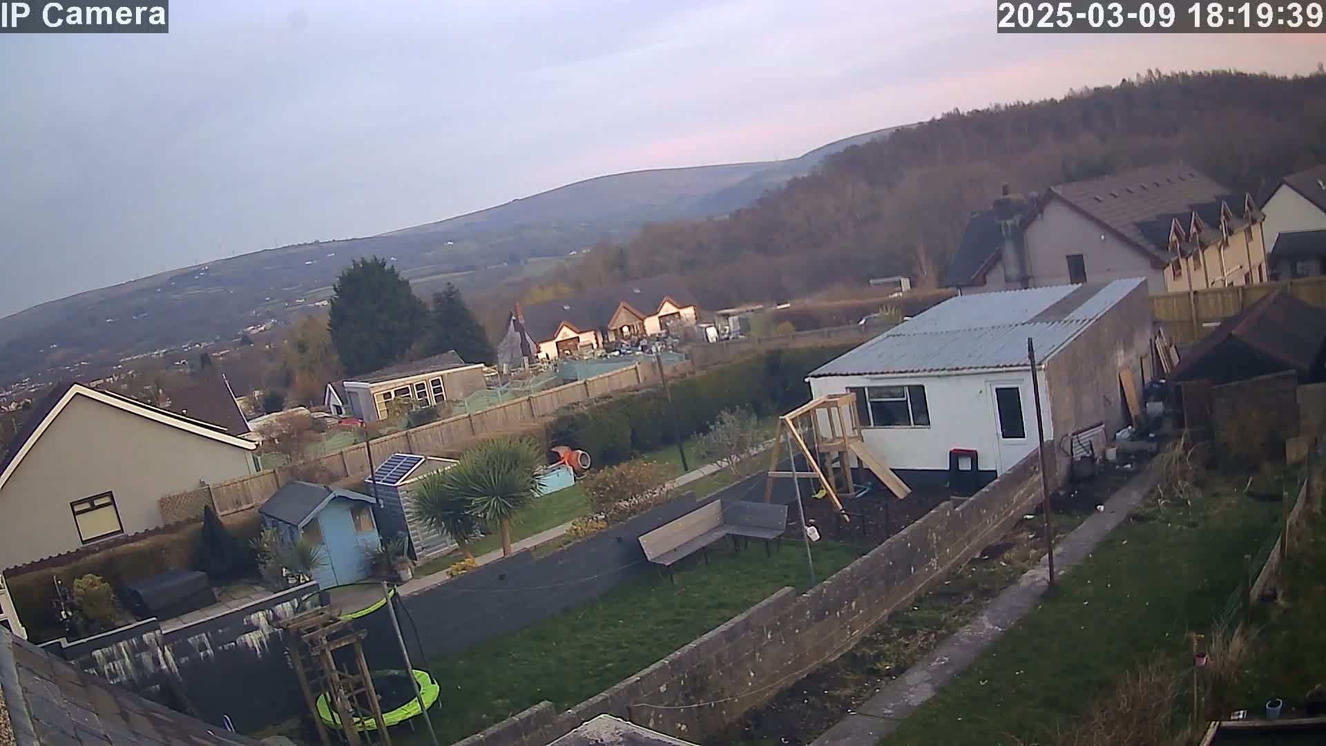 An overcast day reveals a backyard scene with houses, gardens, and a playset, overlooking distant hills.