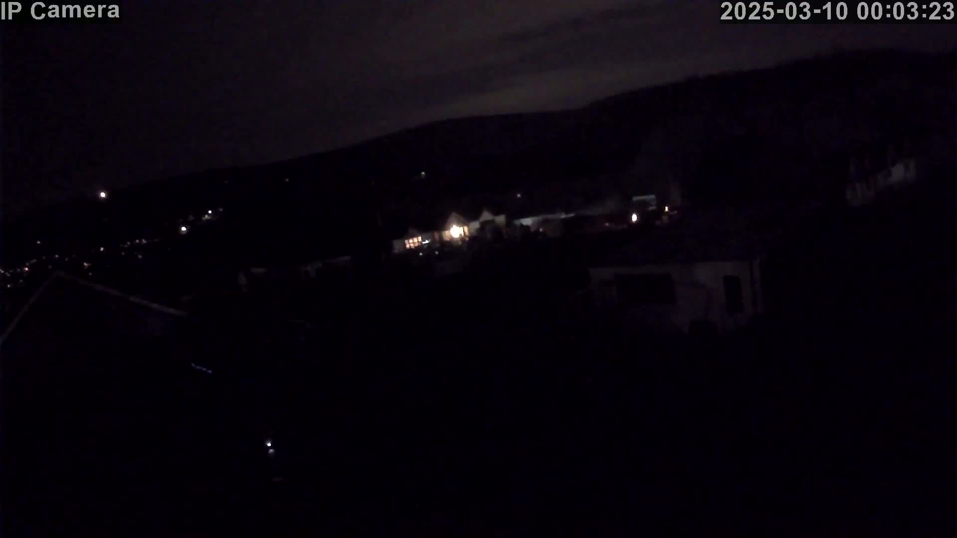 A nighttime, low-light view shows a dimly lit landscape with a few scattered lights and a dark, hilly background under a cloudy sky.