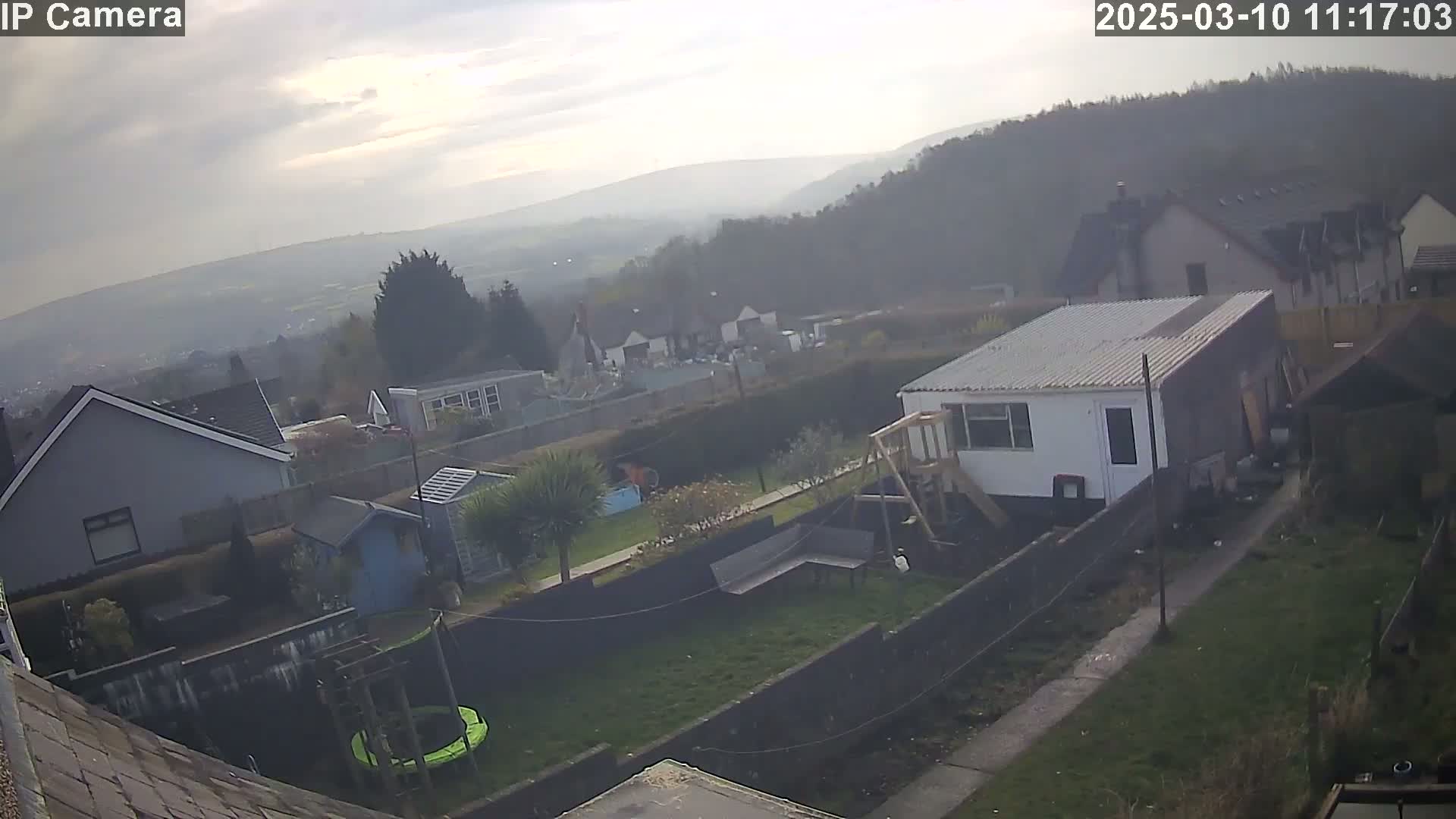 A backyard with houses and a hazy view of distant hills under a partly cloudy sky.