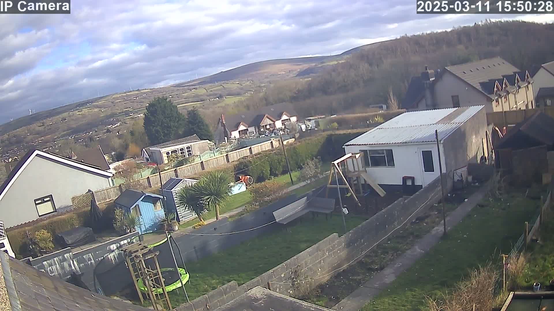 A backyard scene on a partly sunny day shows a residential area with houses and gardens, including a trampoline, sheds, and a playset, all overlooking a valley and distant hills.