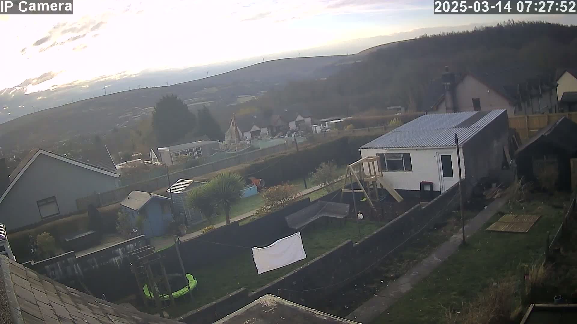 A hazy sunrise illuminates a residential backyard with a small white building, a trampoline, and laundry hanging on a line, overlooking a valley dotted with wind turbines.