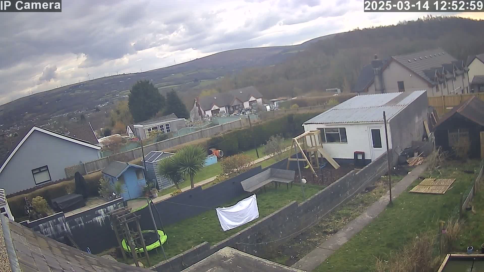A backyard scene under a cloudy sky shows houses and a garden with a trampoline, shed, and play structure, all overlooking a valley with wind turbines in the distance.