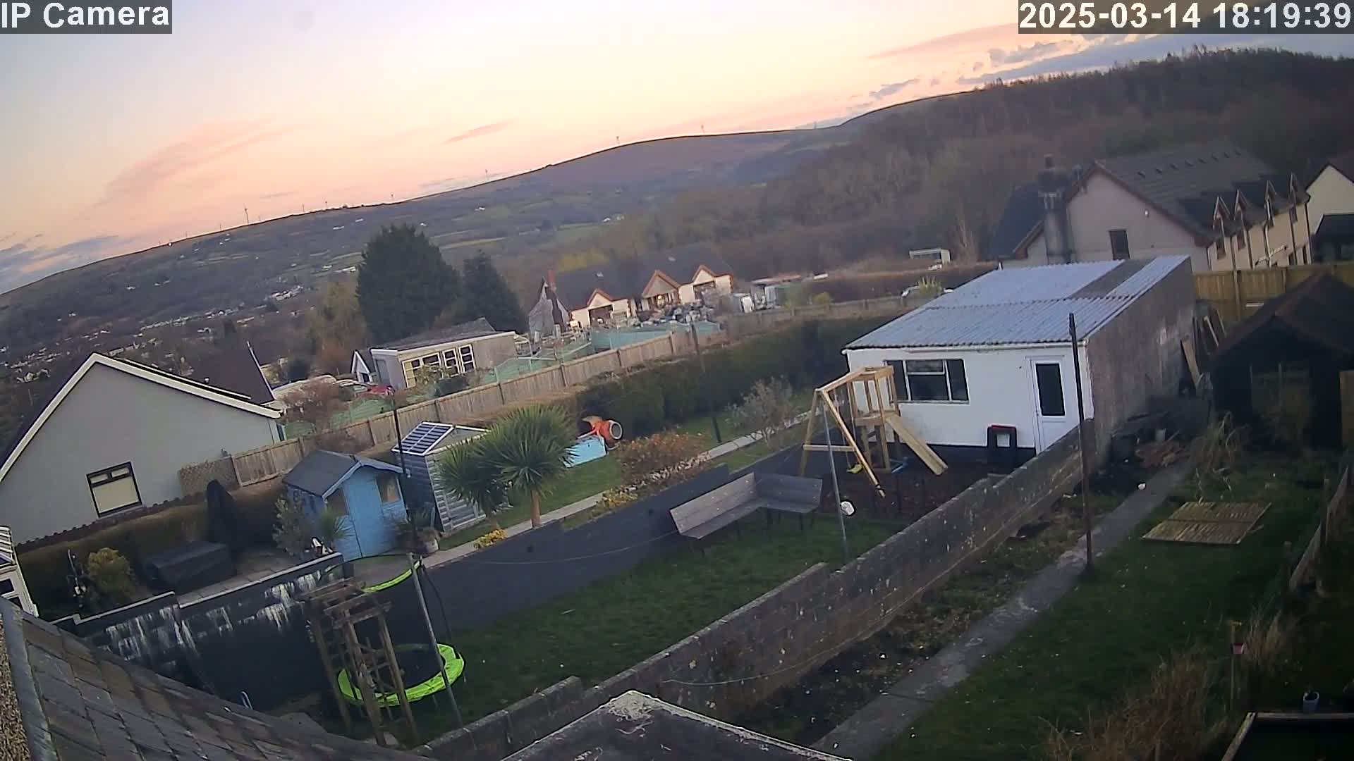A backyard scene at sunset shows a small building, playground equipment, garden shed, and trampoline, with a view of houses and hills in the background under a partly cloudy sky.