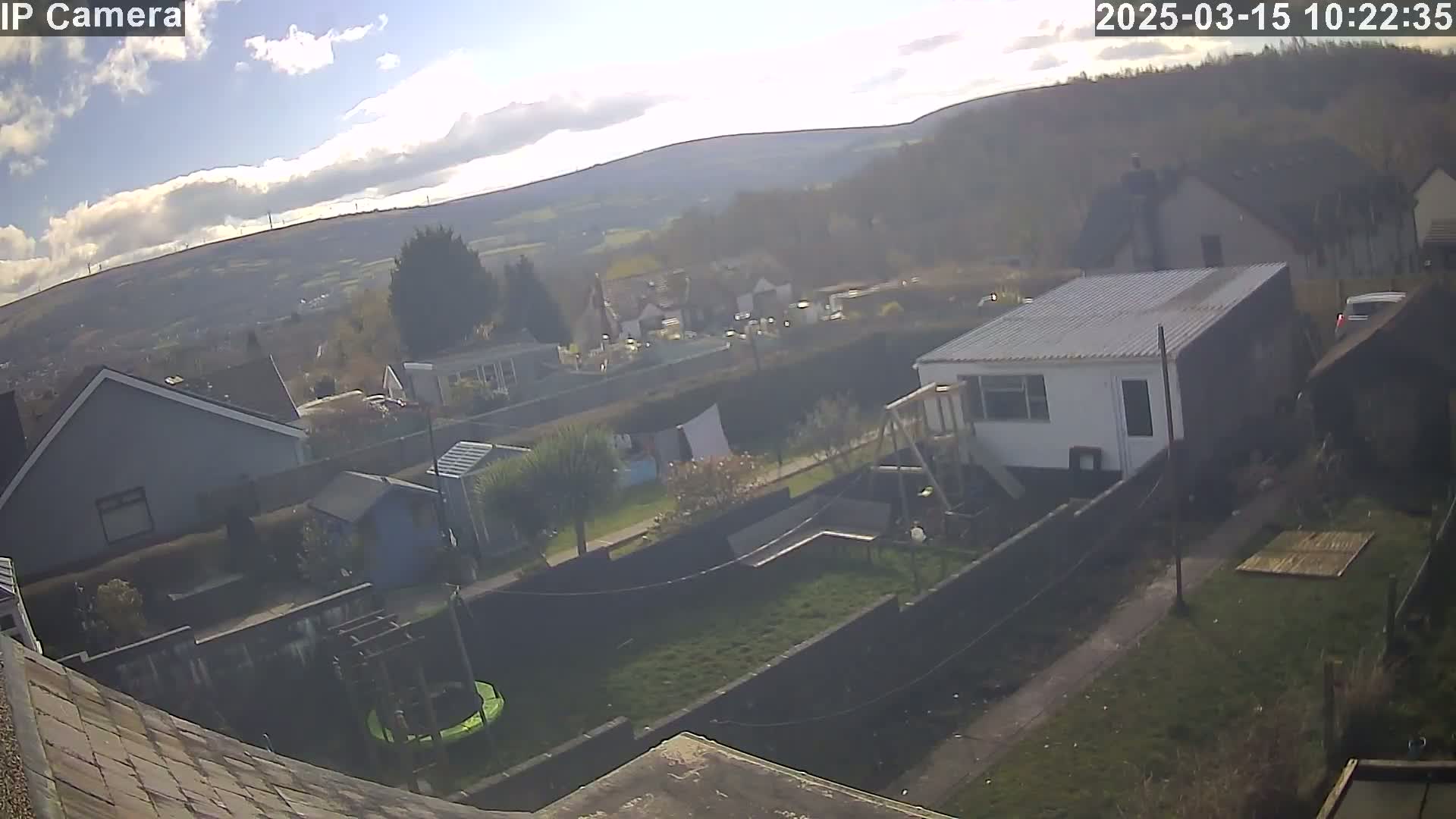 A sunny day reveals a backyard scene with houses, a garden, a trampoline, and distant hills under a partly cloudy sky.