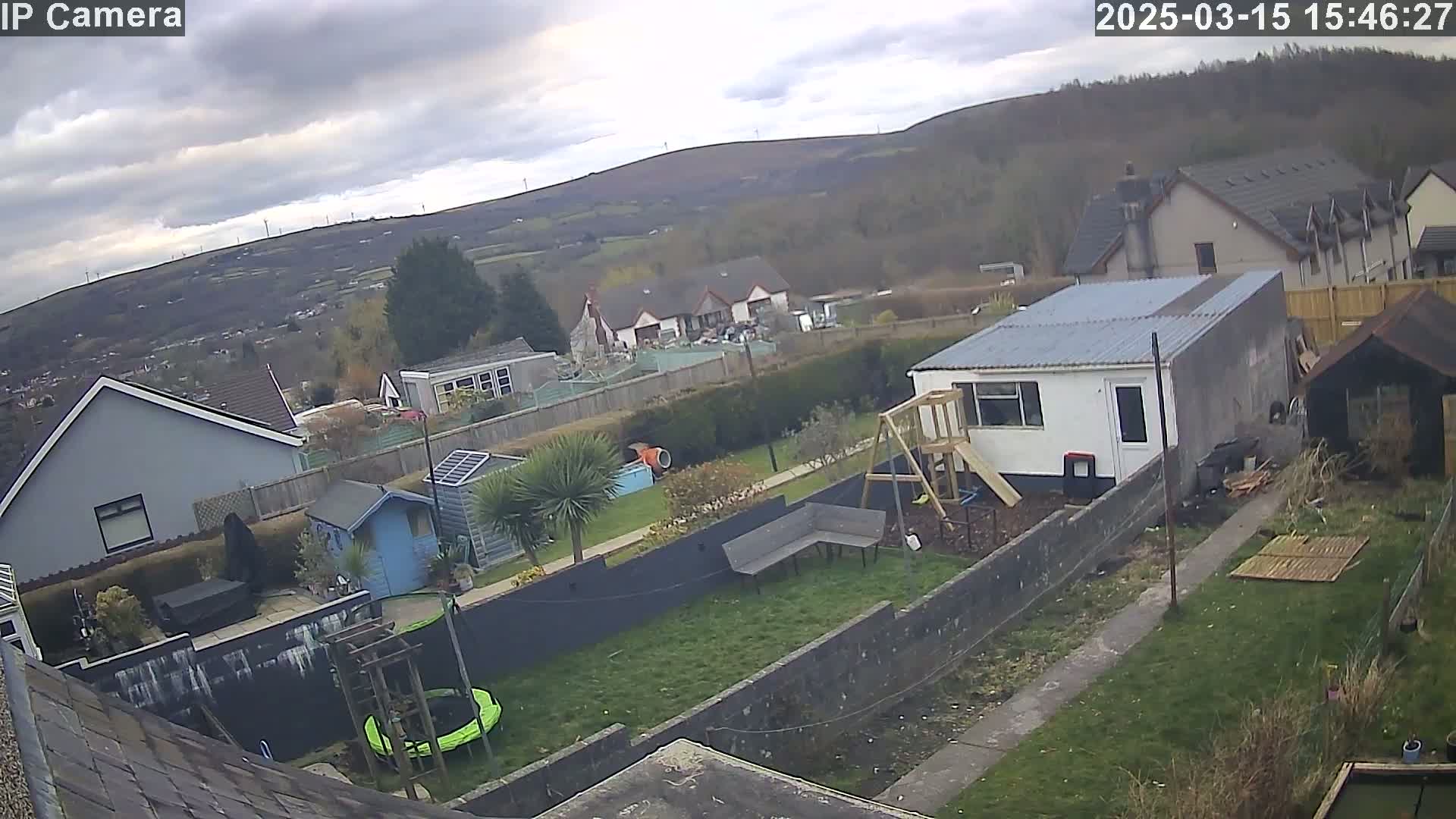A backyard scene on an overcast day features a garden with a trampoline, playset, and small buildings, overlooking houses and hills in the distance.