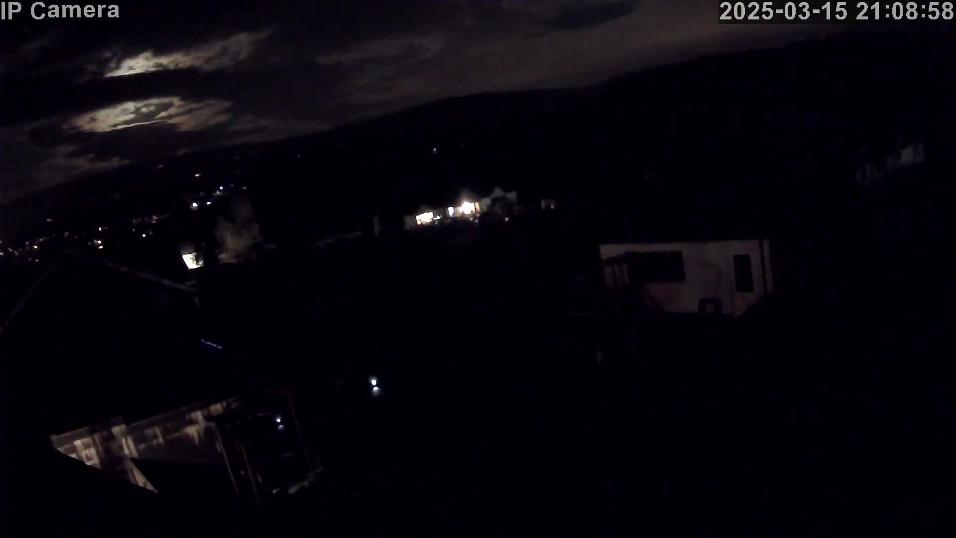 A nighttime, high-angle view shows a dark landscape with a few scattered lights from buildings under a partly cloudy sky.