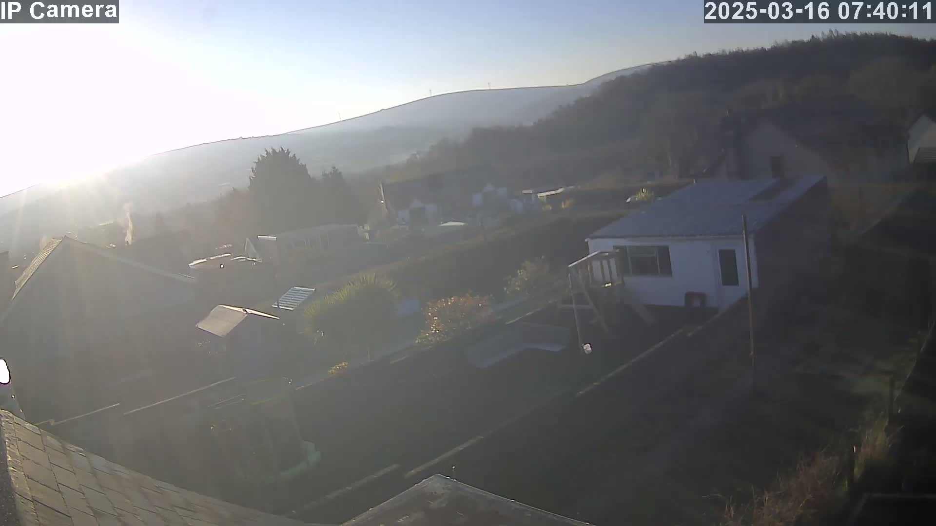 A hazy sunrise illuminates a residential area nestled against a hillside, showcasing houses and gardens.