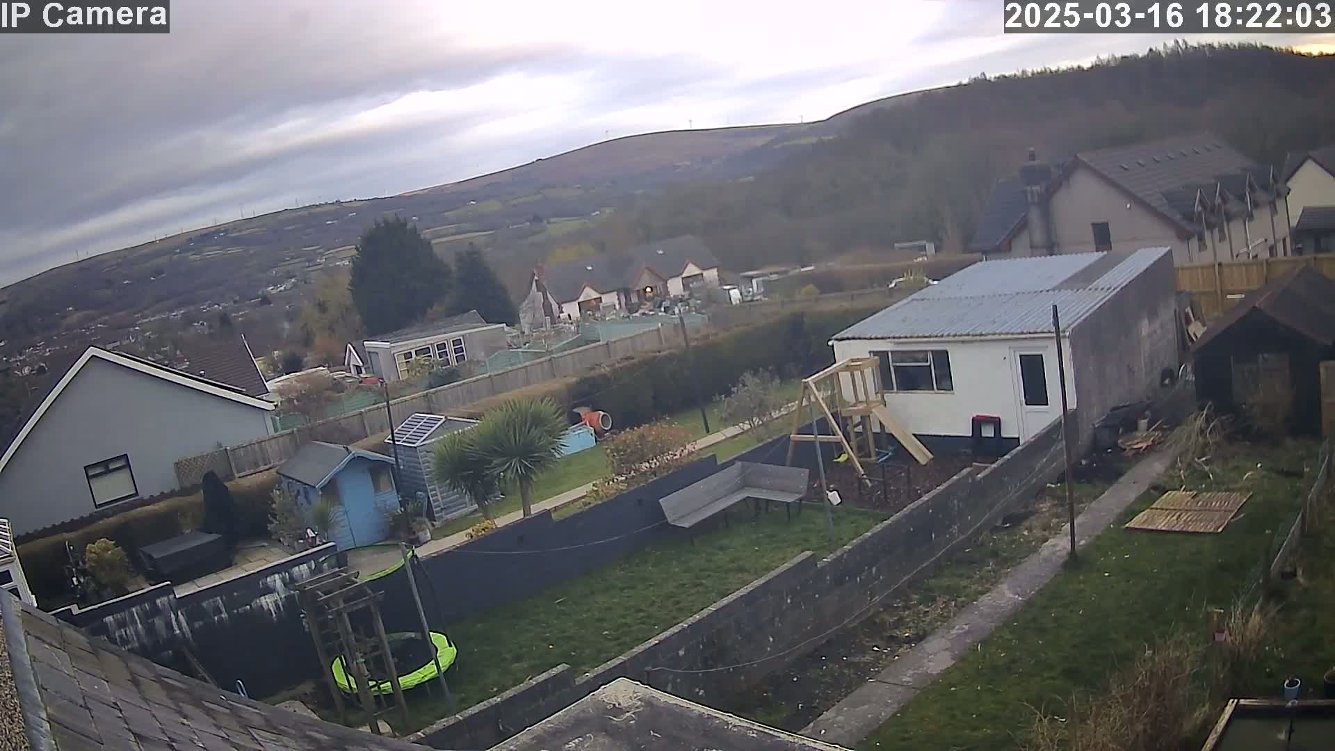 An overcast day reveals a backyard scene with houses and hills in the distance, featuring a trampoline, a playset, and a small shed.