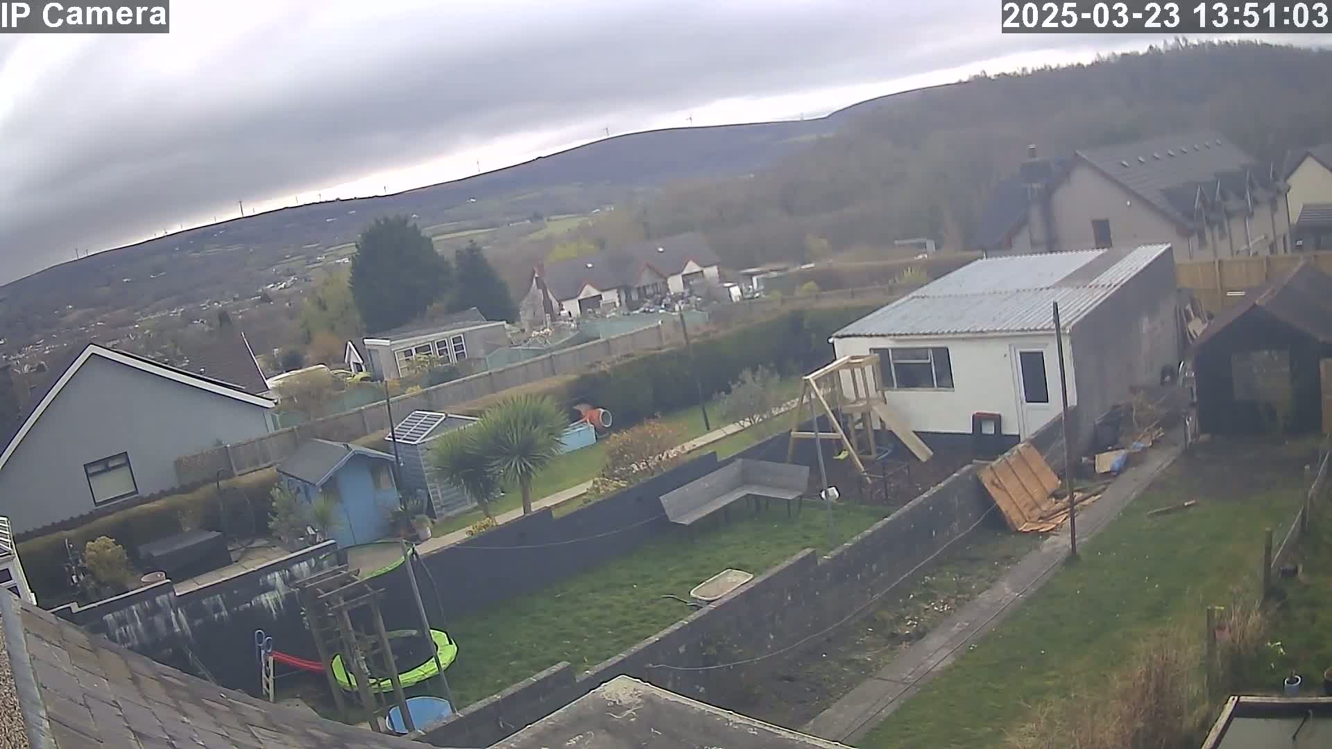 An overcast day reveals a backyard scene with houses, a garden, play structures, and a shed, all set against a backdrop of distant hills.