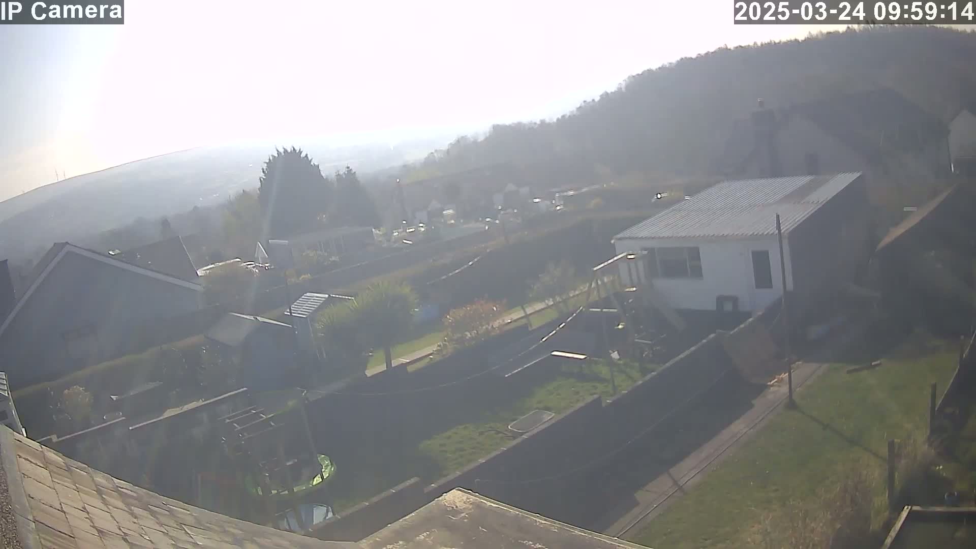 A hazy, sunny view of a residential area, showing houses, gardens, and a small building, with hills visible in the background.
