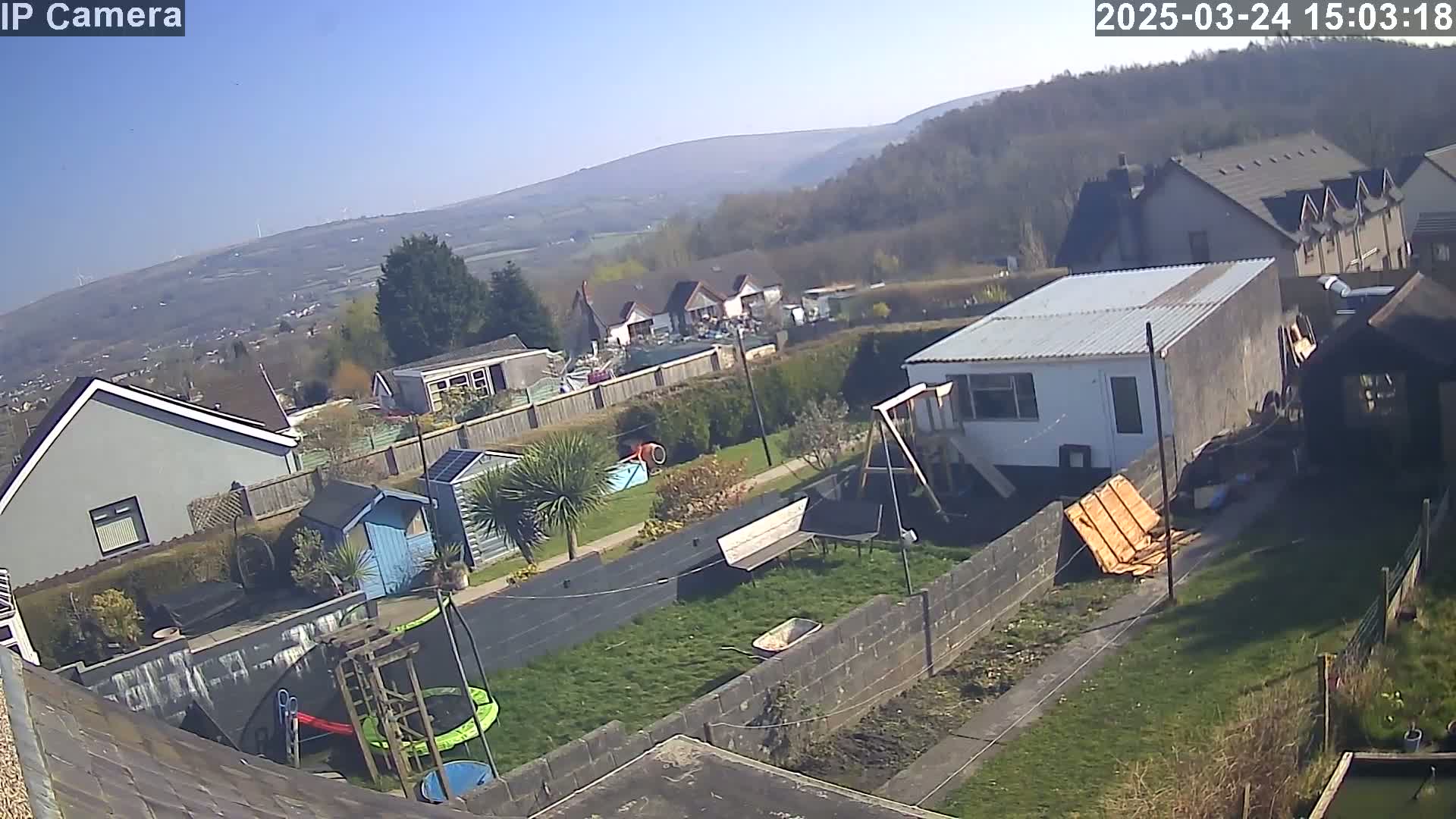 A backyard scene on a sunny day features houses, a small garden with a trampoline and playset, and a view of distant hills.
