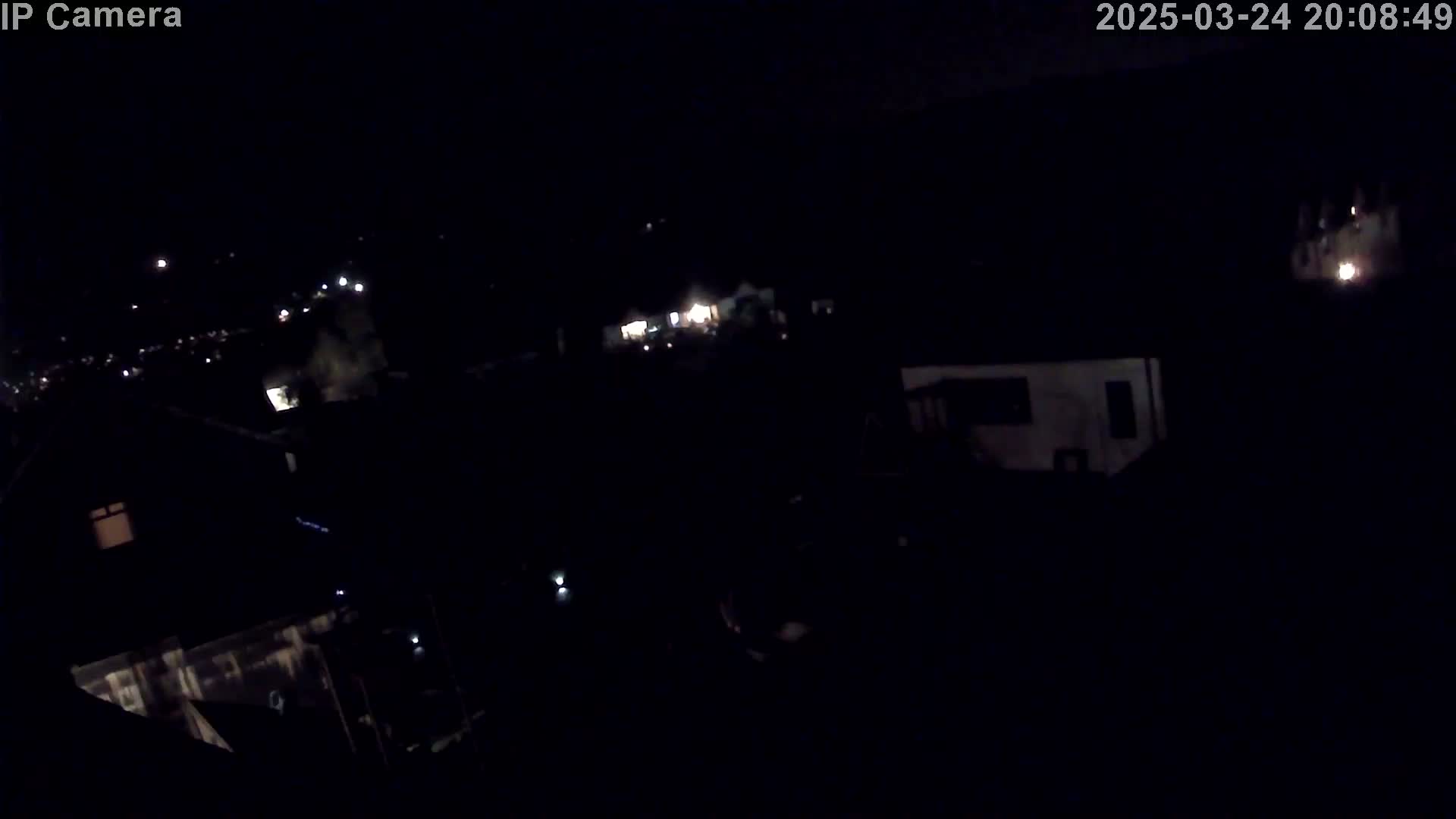 A nighttime, high-angle view shows dimly lit houses and a few bright lights in the distance under a dark sky.