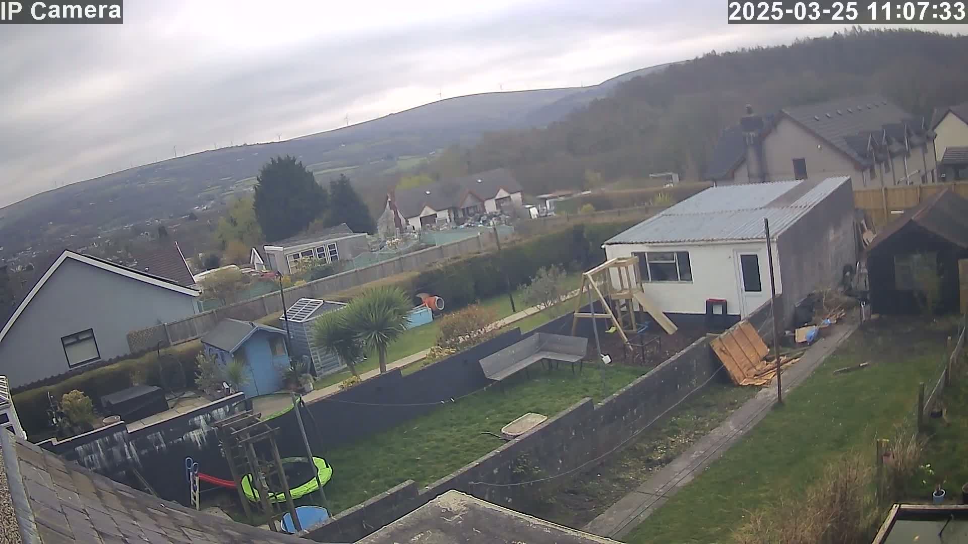 An overcast day reveals a backyard scene with houses and hills in the distance, featuring a playset, trampoline, and small outbuildings.