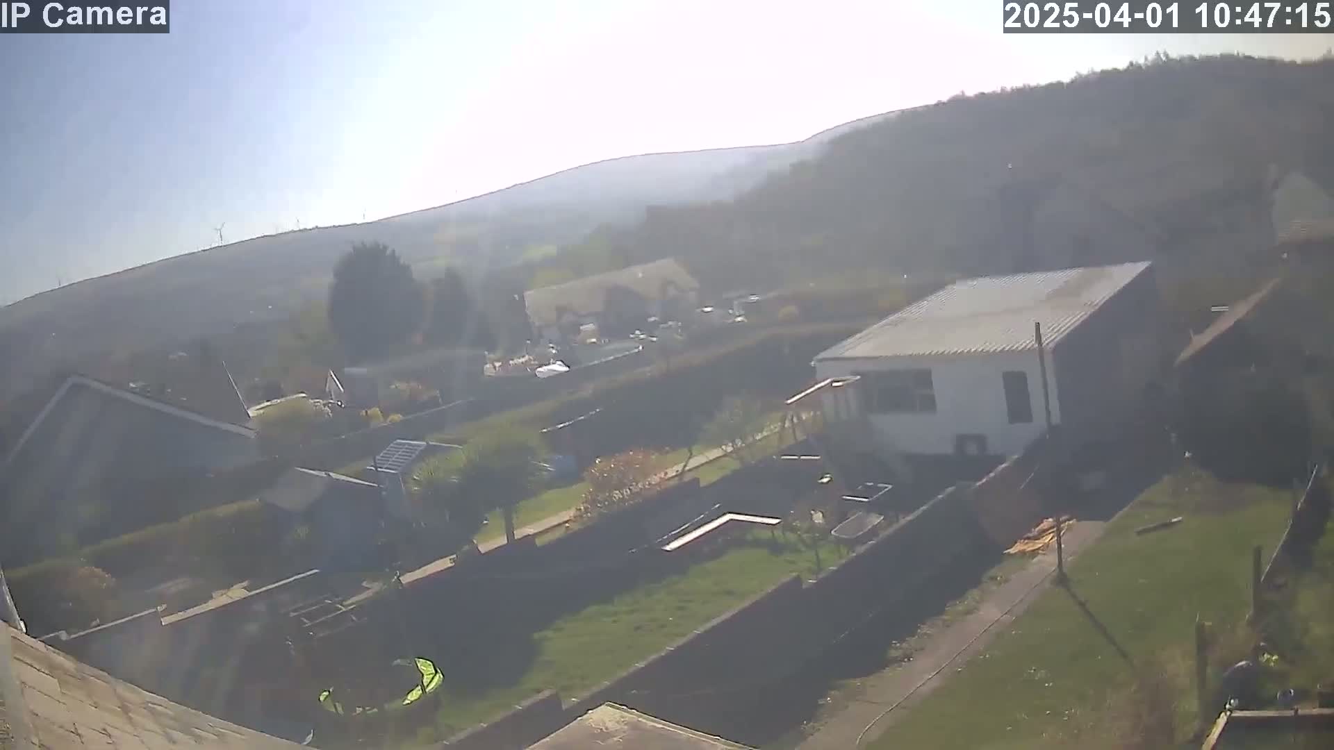 A high-angle, long shot on a sunny day shows a residential backyard with a small white building, a grassy area, and houses and hills in the background.