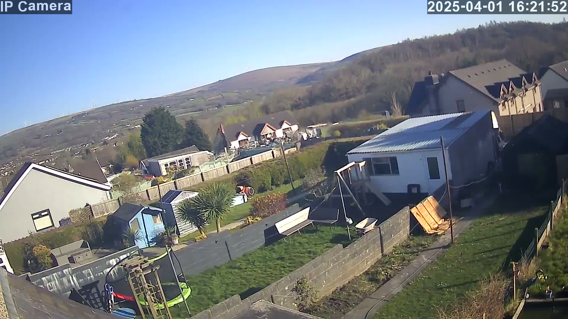 A sunny day overlooks a residential backyard with a mobile home, garden, and playground equipment, situated on a hillside with a distant view of more houses and wind turbines.