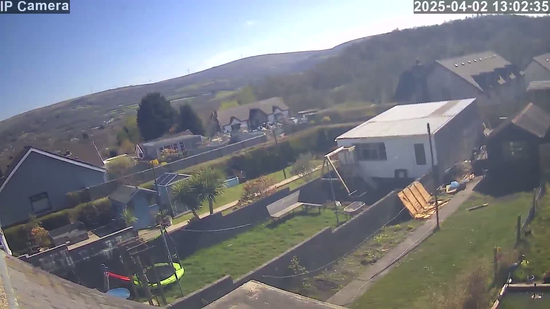 An aerial view shows a residential backyard with a green lawn, several buildings, and a sunny day with clear blue skies and distant hills.