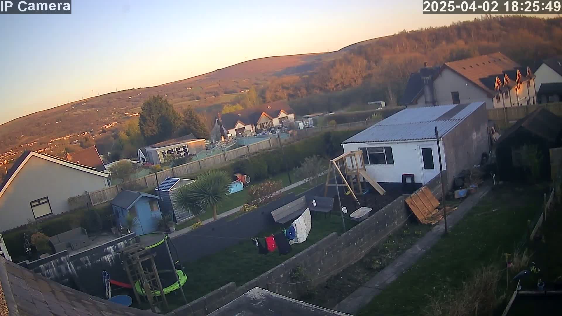 An aerial view of a residential backyard with a trampoline, playset, and clothesline, overlooking a neighborhood nestled against a hill at sunset.