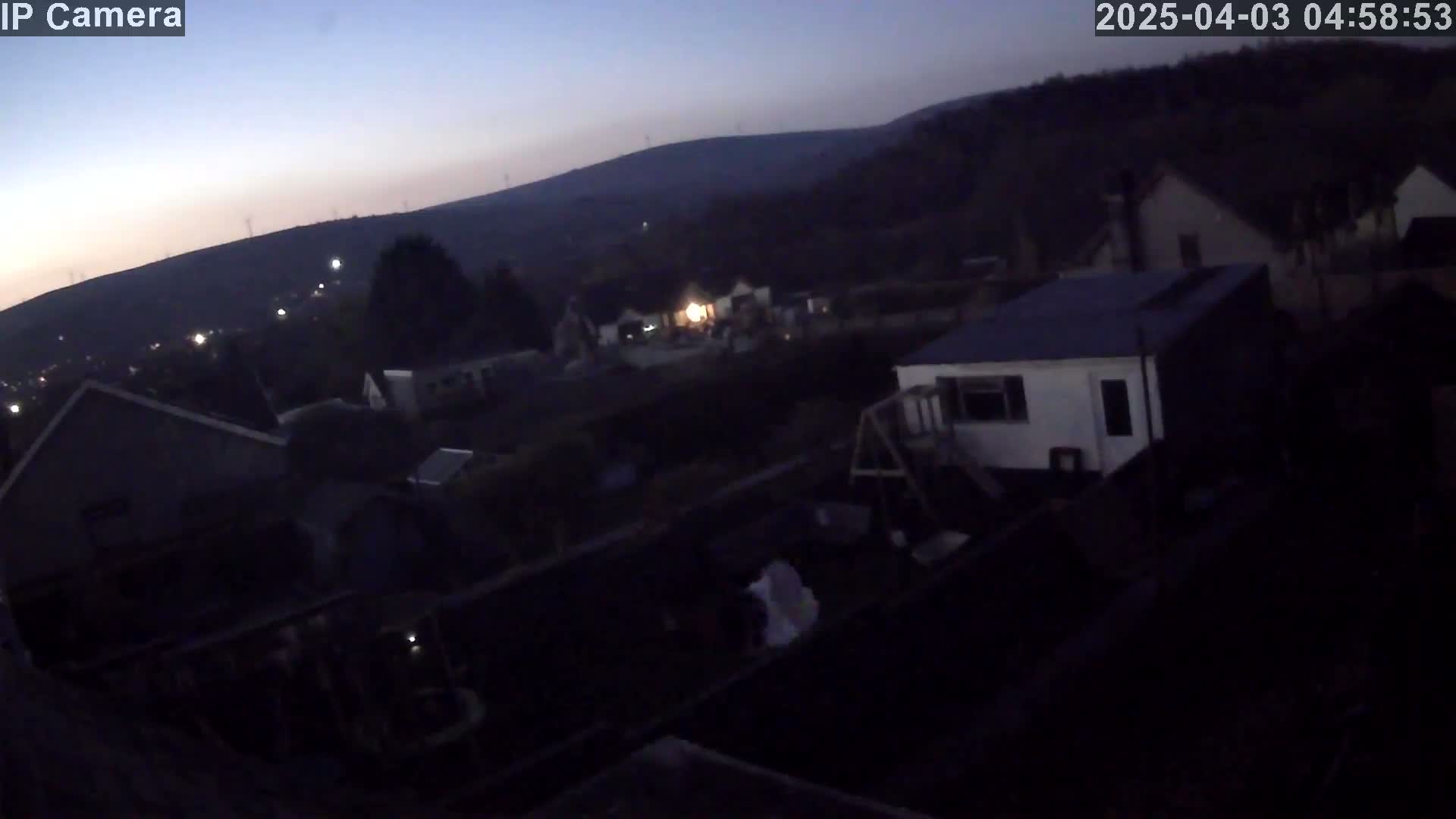 A dark, nighttime view of a residential area with houses and a hilly landscape in the background, under a twilight sky.