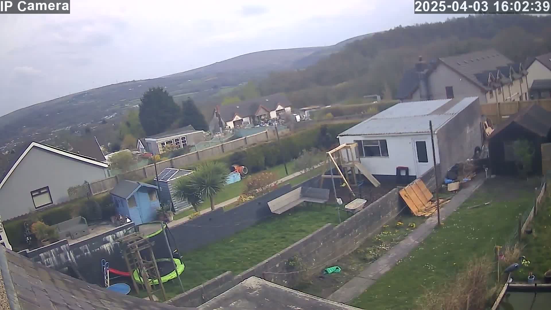 An overcast day reveals a backyard scene with houses and hills in the distance, featuring a trampoline, playset, and garden.