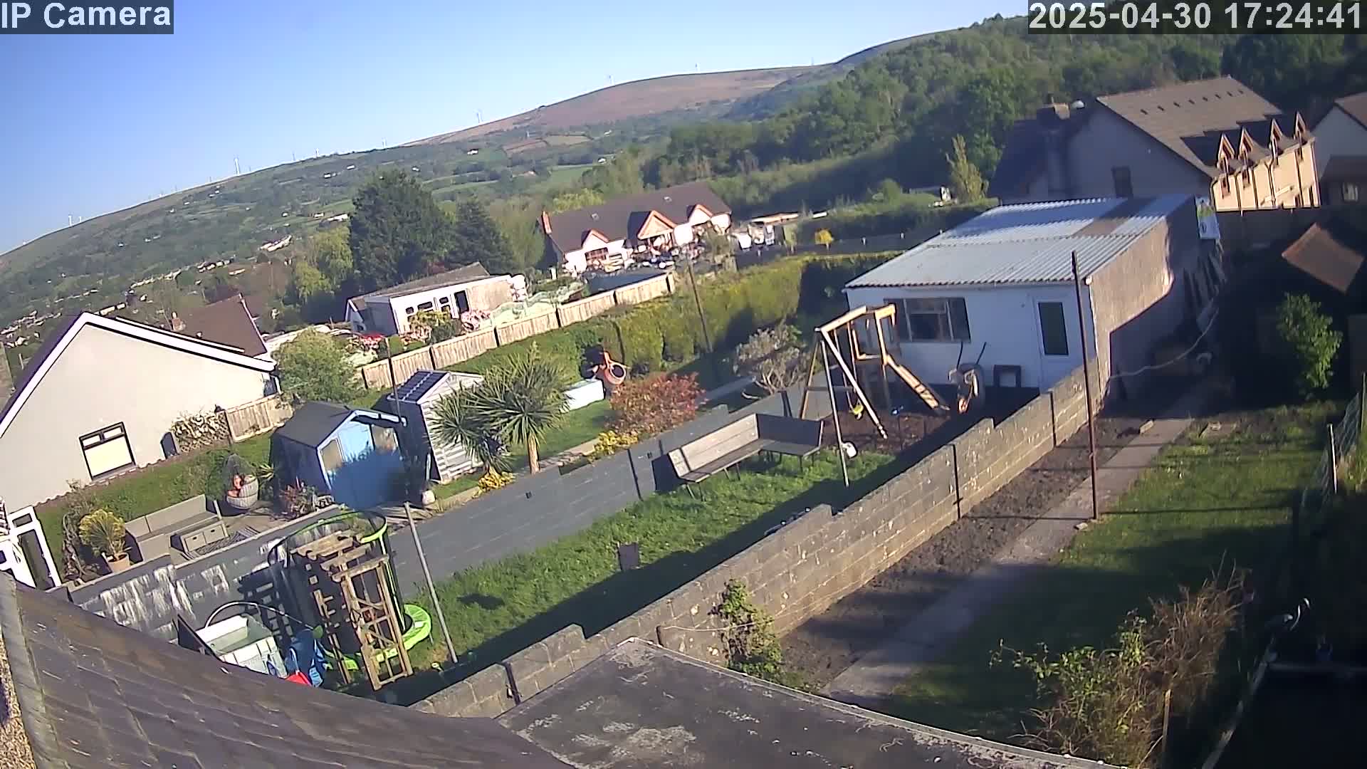 An aerial view shows a sunny residential area with houses, gardens containing sheds, play equipment, and a stone wall, all situated below a green hill.