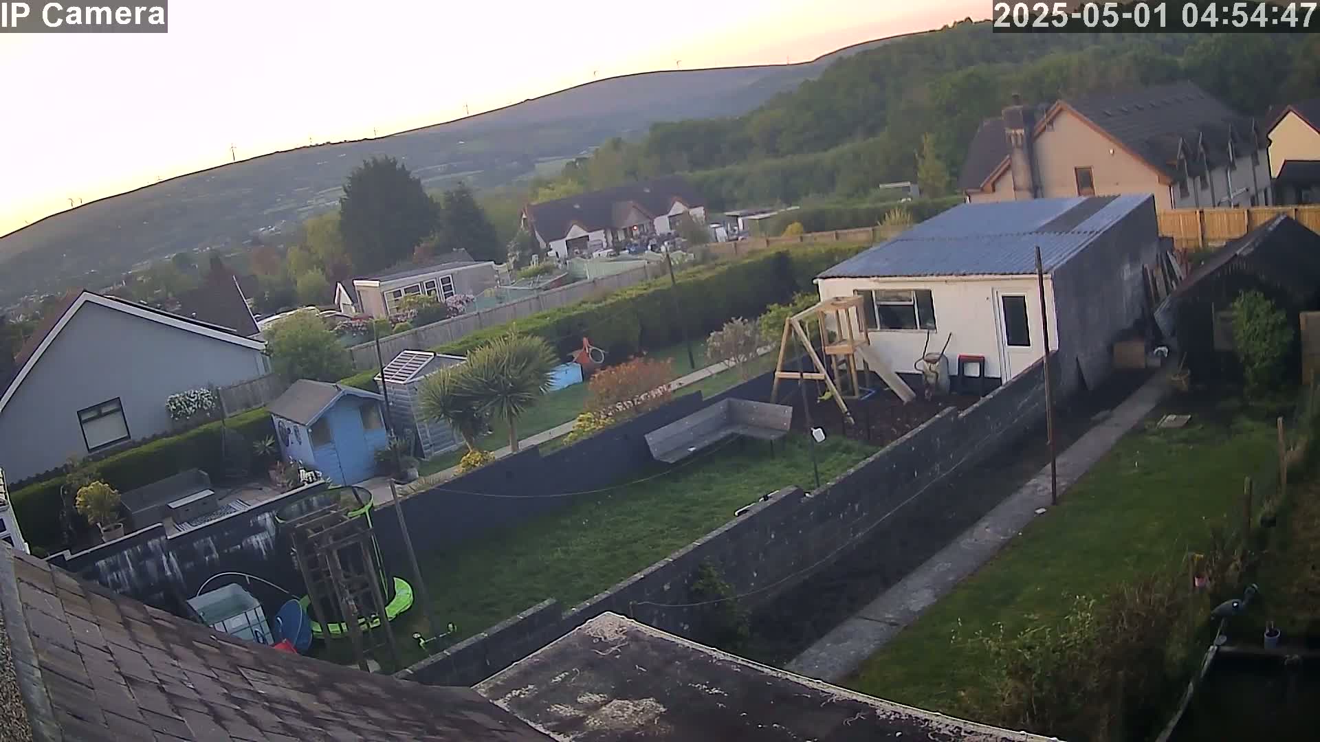 A high-angle view shows a backyard with a small building, a swing set, and a lawn, all set against a backdrop of houses and rolling hills under a twilight sky.