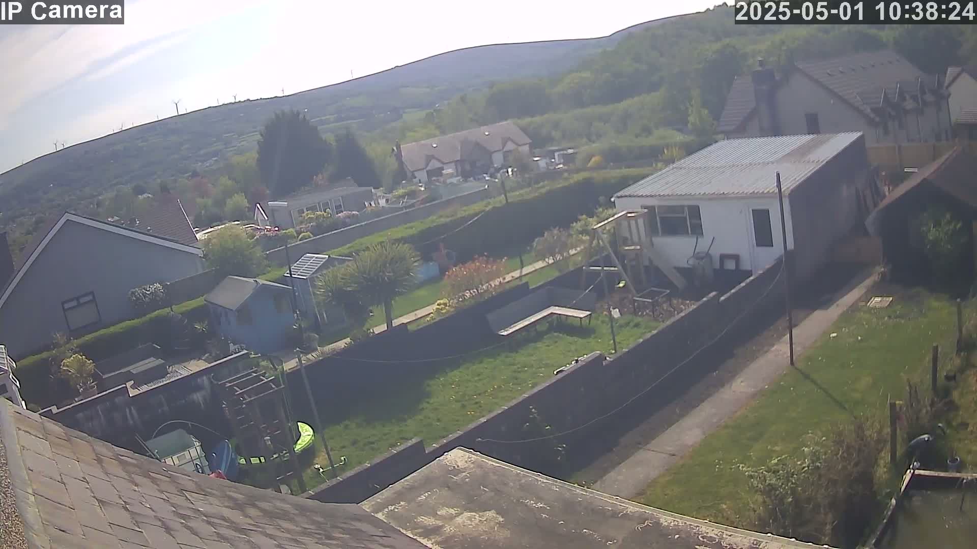 A hazy, sunny day overlooks a residential area with houses, gardens, and a mobile home, all situated against a backdrop of distant hills and wind turbines.