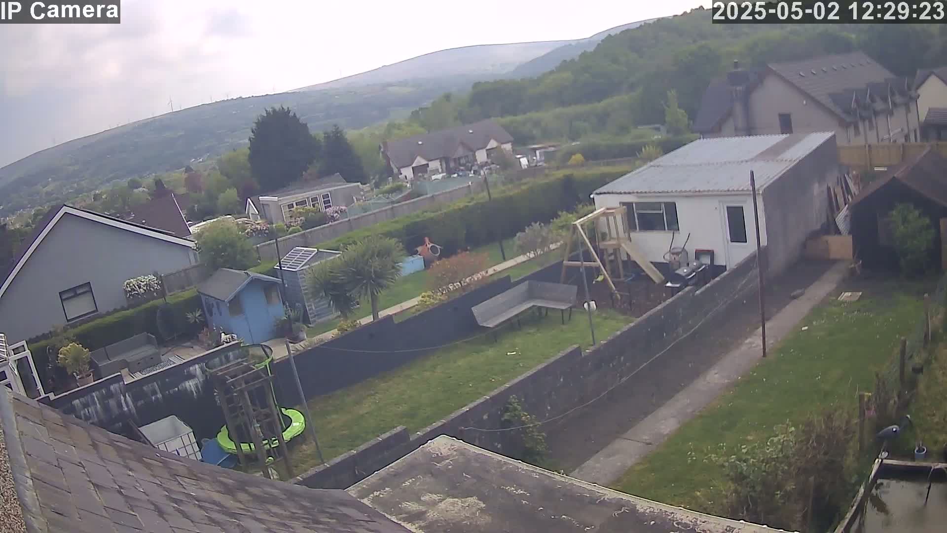An overcast day reveals a backyard scene with houses and hills in the background, a garden with a shed, playset, and a pond.