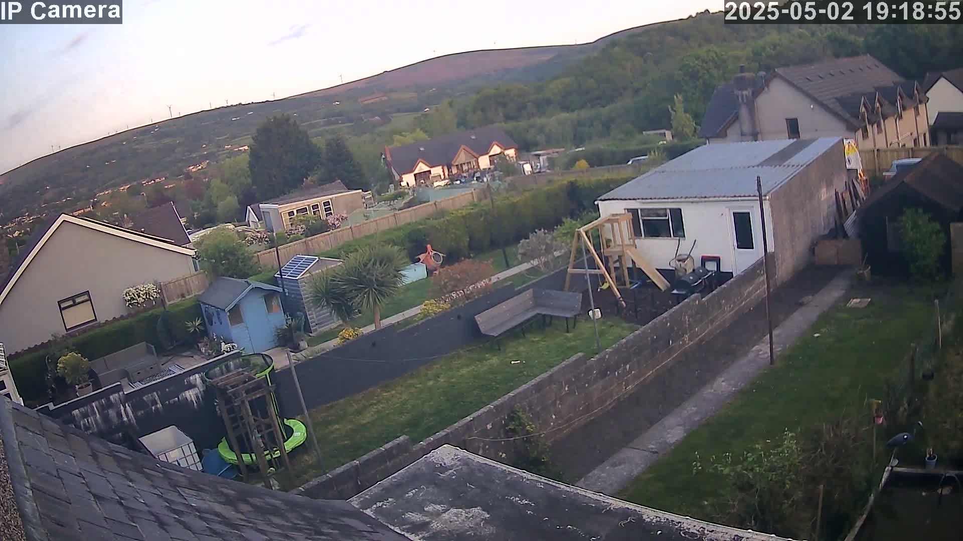 An aerial view of a residential backyard at dusk, showing houses, gardens, and a playset, with a hill visible in the background.
