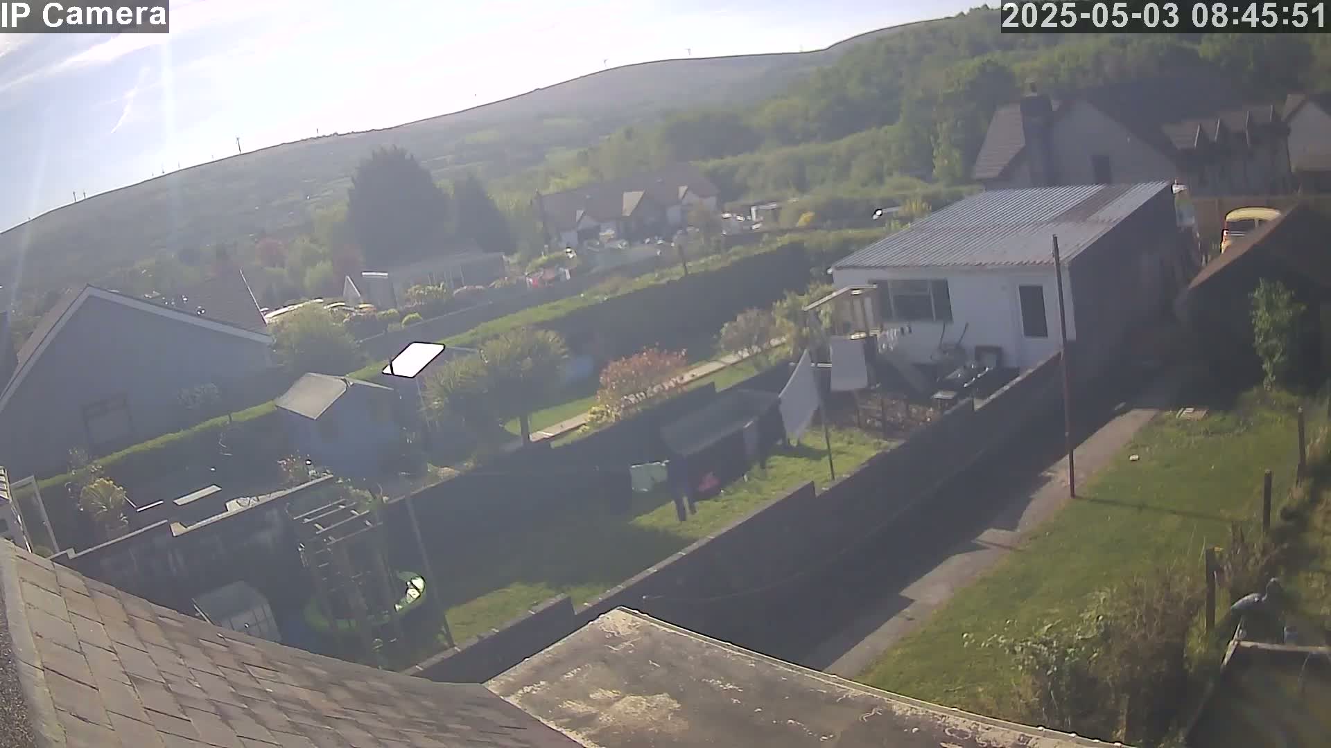 A hazy, sunny view overlooks a suburban residential area with houses, gardens, and a small shed, all surrounded by green hills in the background.