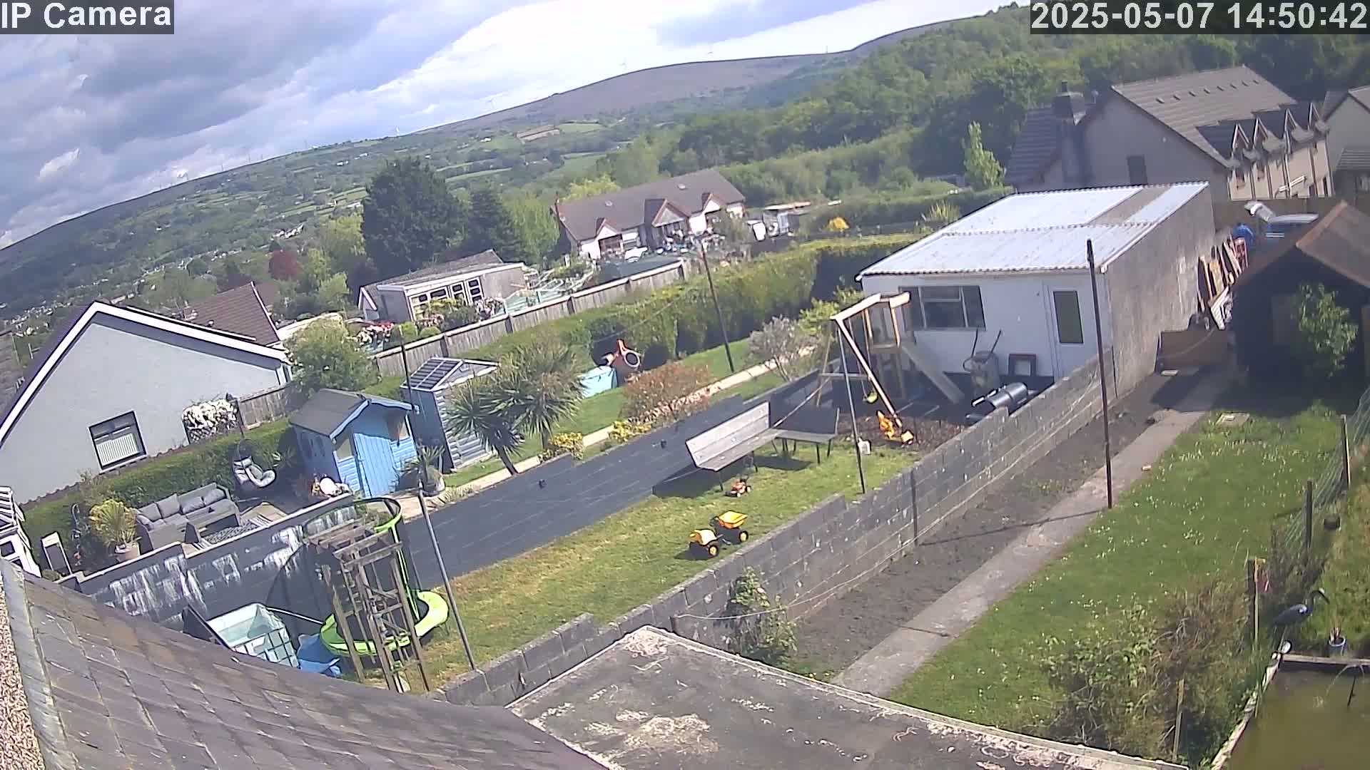 A partly cloudy day overlooking a residential backyard with houses and greenery, including a small pond and play equipment, under a mostly cloudy sky.