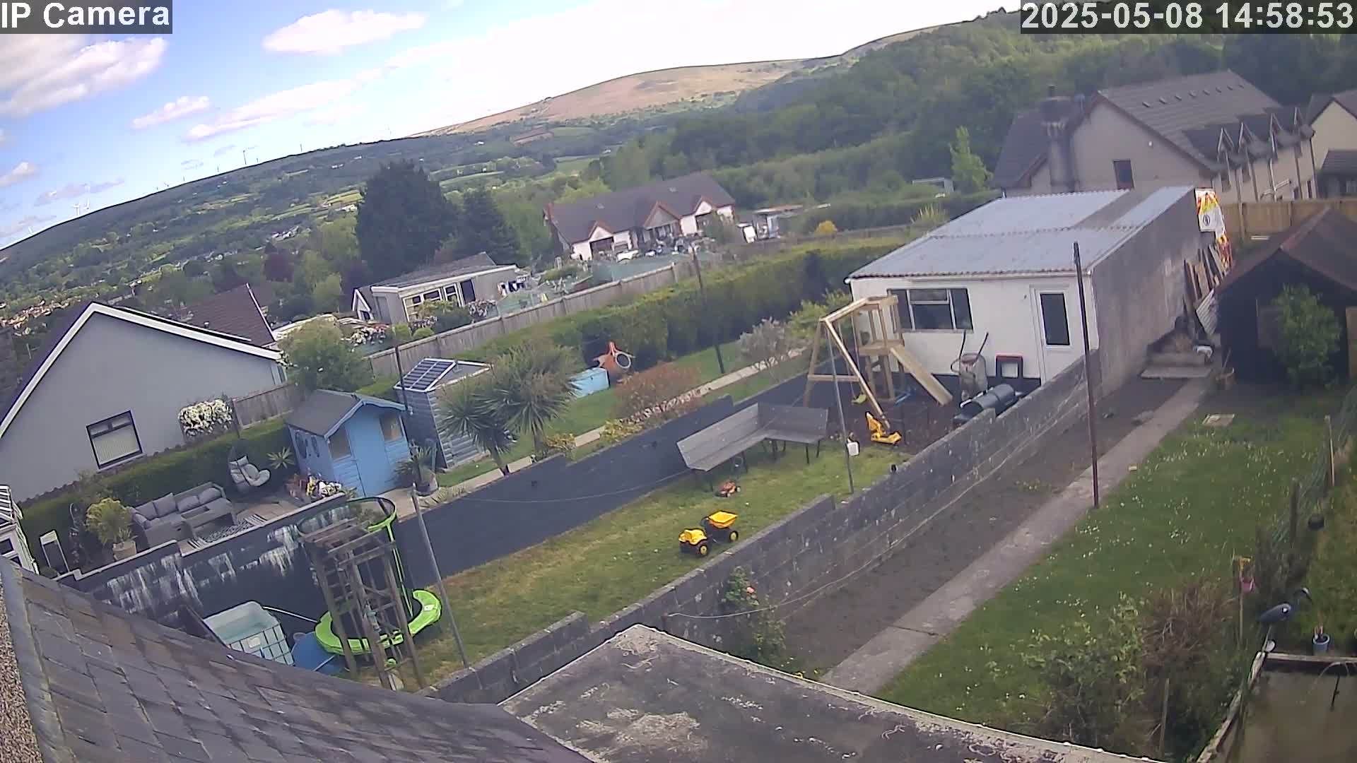 A backyard scene on a partly cloudy day shows a house, a garden with lawn, toys, and a shed, and a view of distant hills and wind turbines.