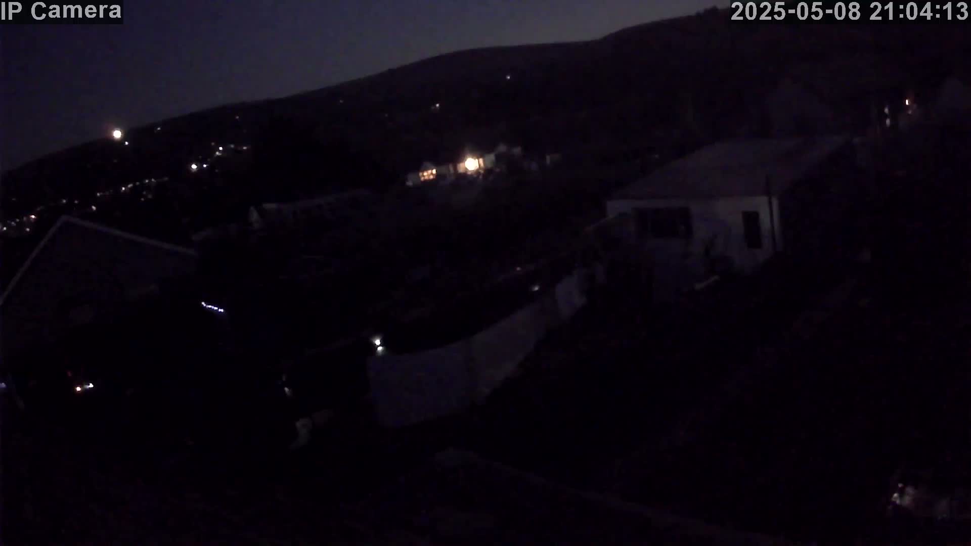 A nighttime, low-light view shows several dark buildings and sparsely lit areas on a hillside under a dark sky.