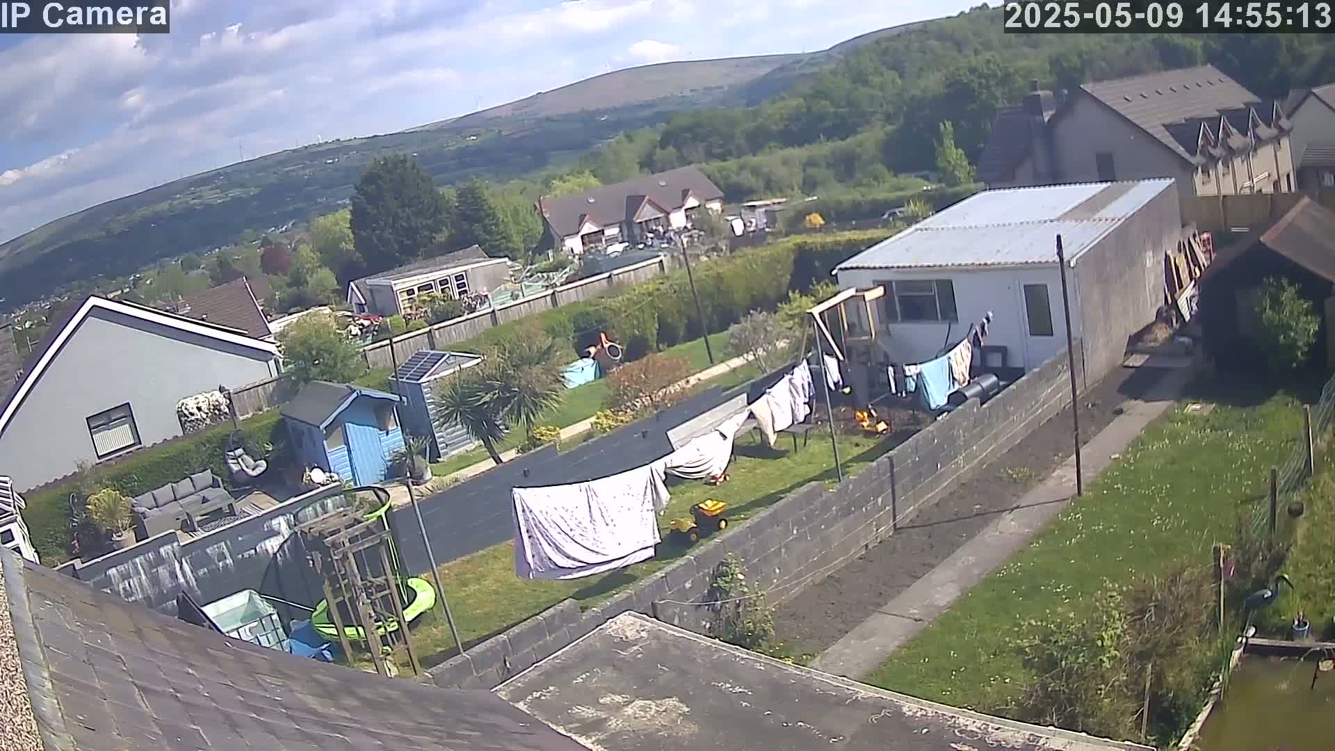 A partly sunny day view of a residential backyard with houses and hills in the background, featuring a clothesline with laundry, a small shed, and a play area.