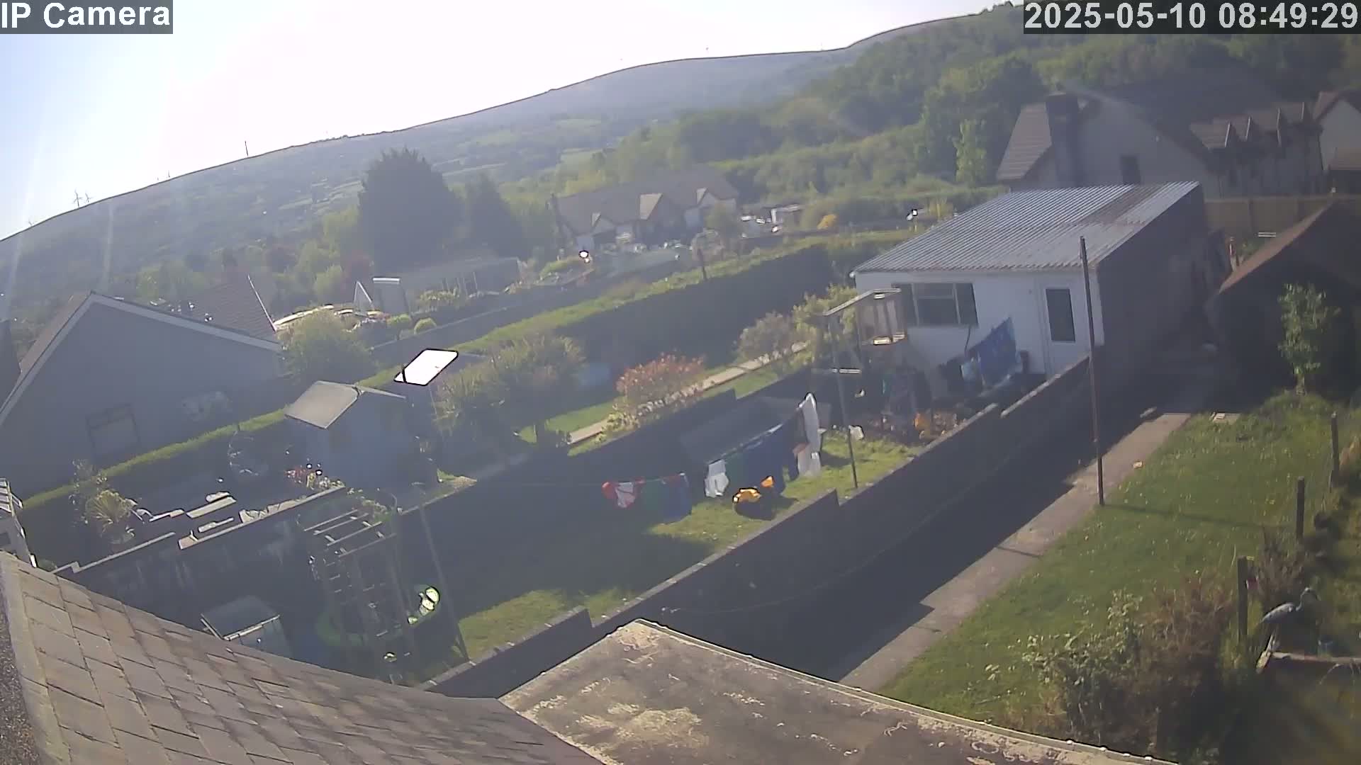 A hazy, sunny day reveals a high-angle view of residential backyards with houses, gardens, and clotheslines, all situated against a backdrop of rolling hills.