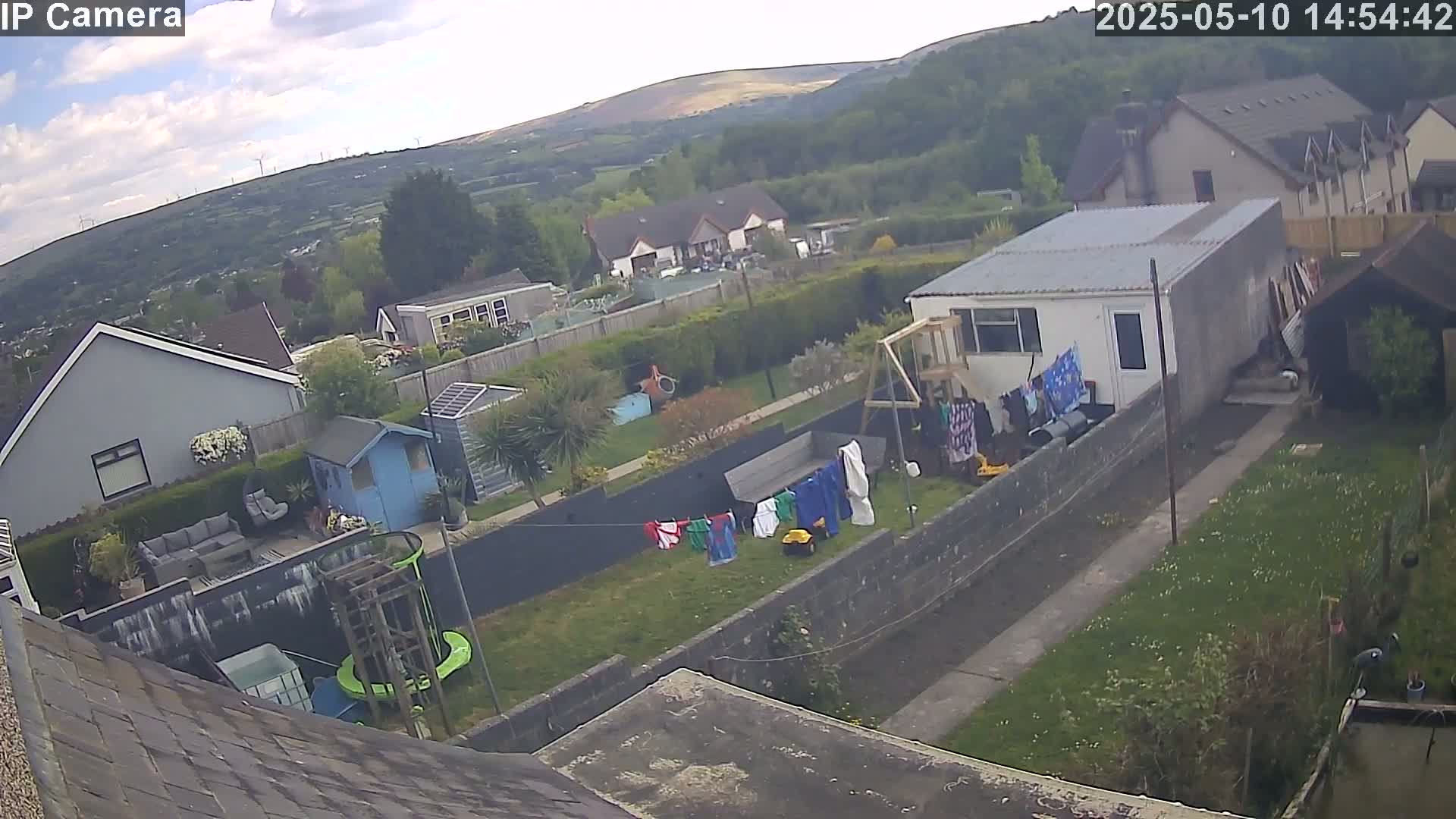 An aerial view of a residential backyard with a partly cloudy sky shows houses and wind turbines in the distance, laundry hanging on a clothesline, and a garden with various structures including sheds and play equipment.