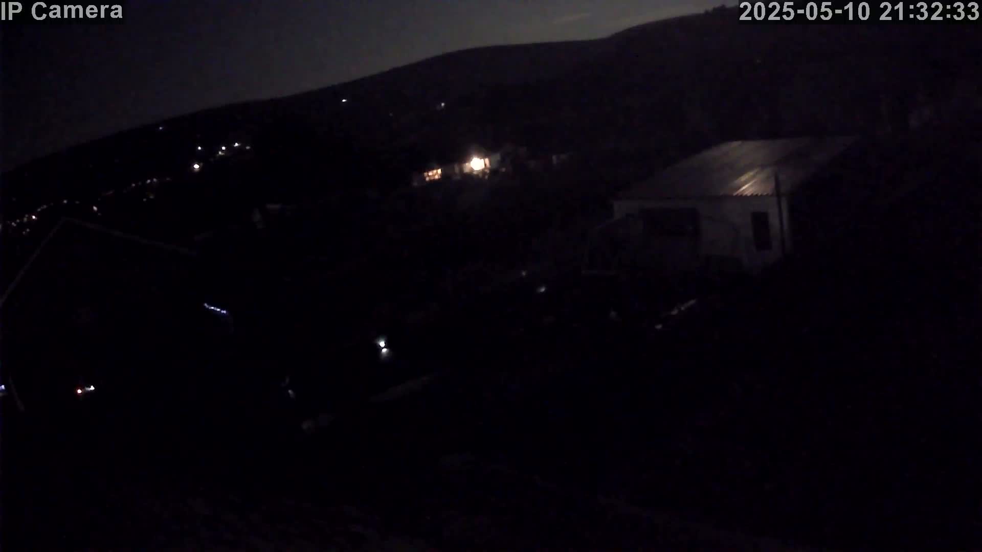 A nighttime, low-light view shows a dark landscape with a few scattered lights and a building in the distance under a mostly clear sky.