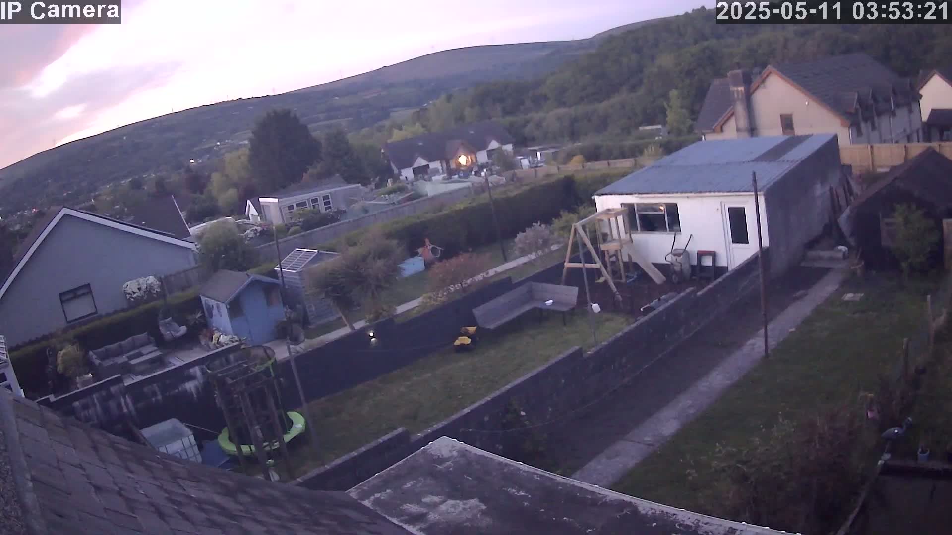 A backyard scene at twilight shows several houses nestled in a hilly, wooded area, with a garden including a playset, shed, and trampoline.