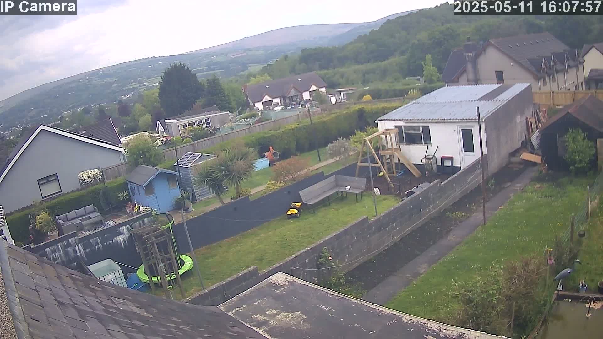 A high-angle view of a backyard with houses and hills in the background under an overcast sky shows a grassy yard with a shed, playset, and garden.