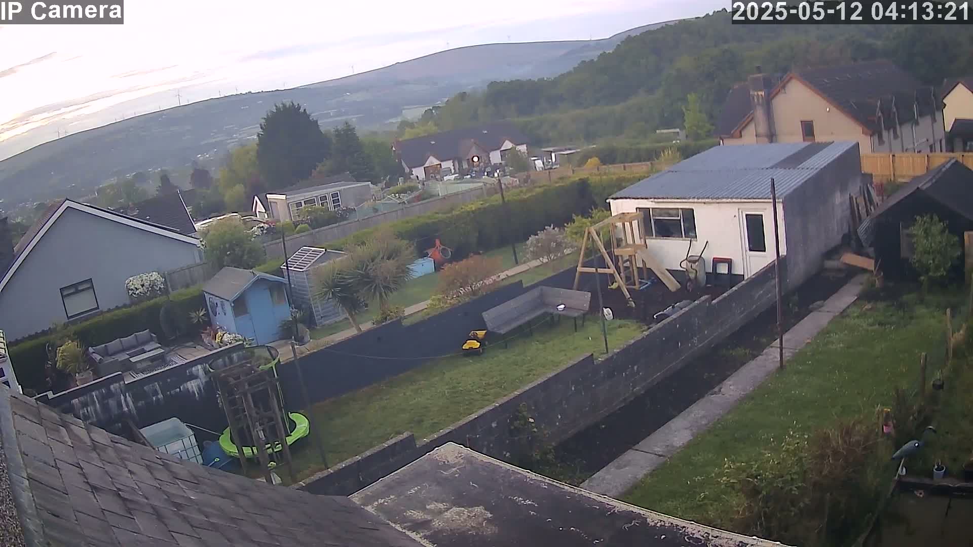 An overcast morning reveals a backyard scene with a small building, a playset, and a lawn, all situated amongst other houses and greenery on a hillside.