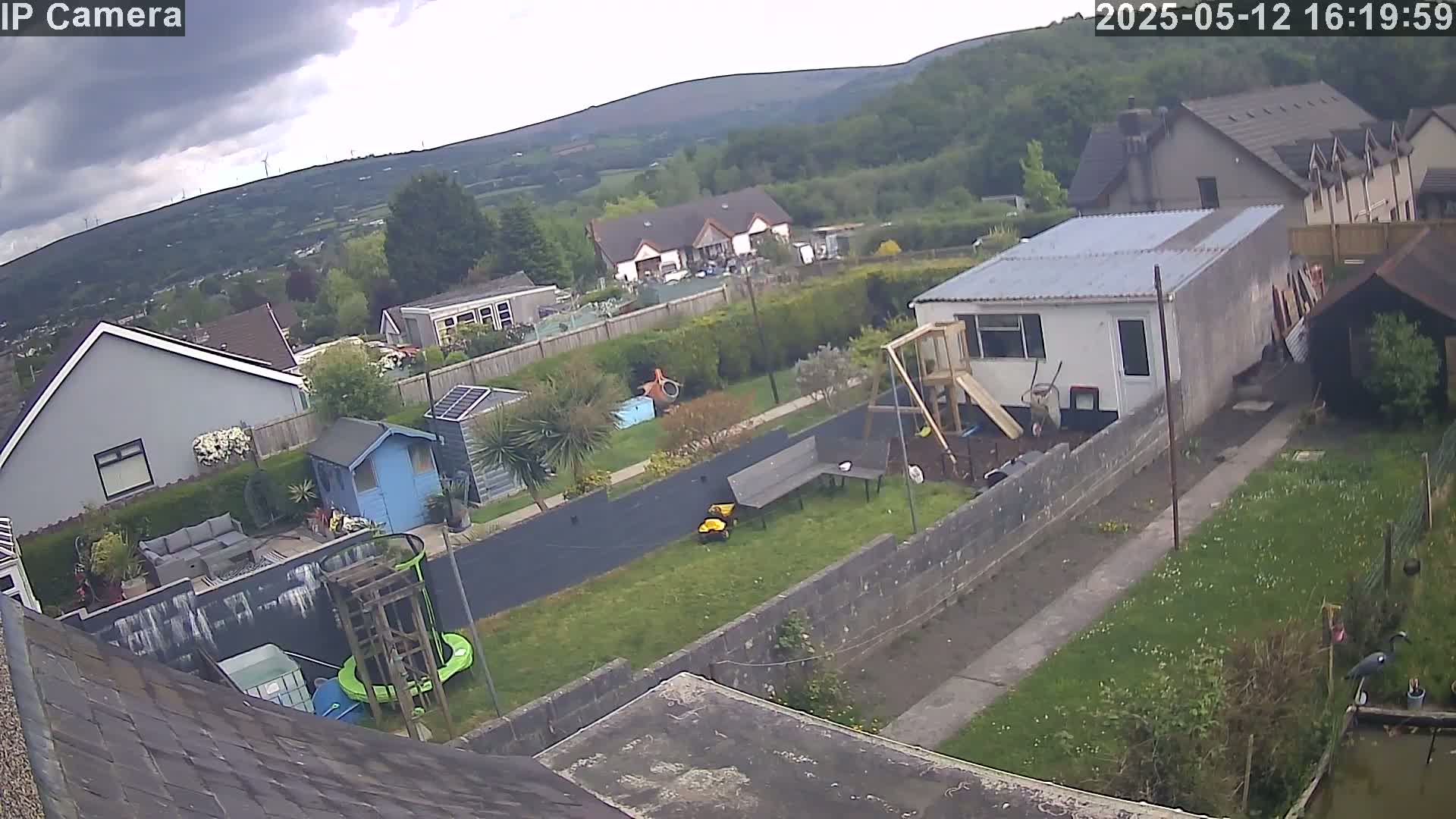 An overcast day shows a backyard with a playset, small shed, and garden, overlooking houses and hills in the distance.