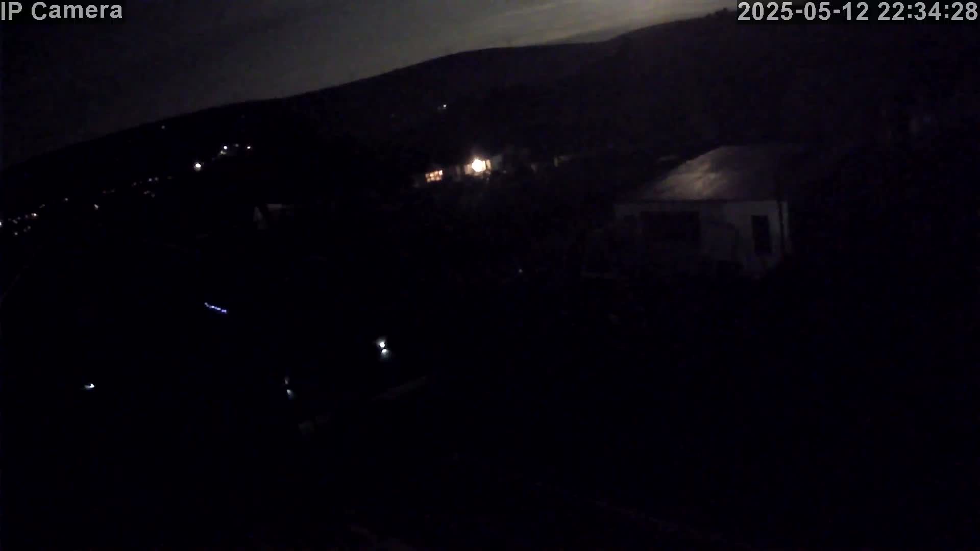 A nighttime, high-angle view shows a dark landscape with a few faint lights and a building visible under a mostly clear sky.