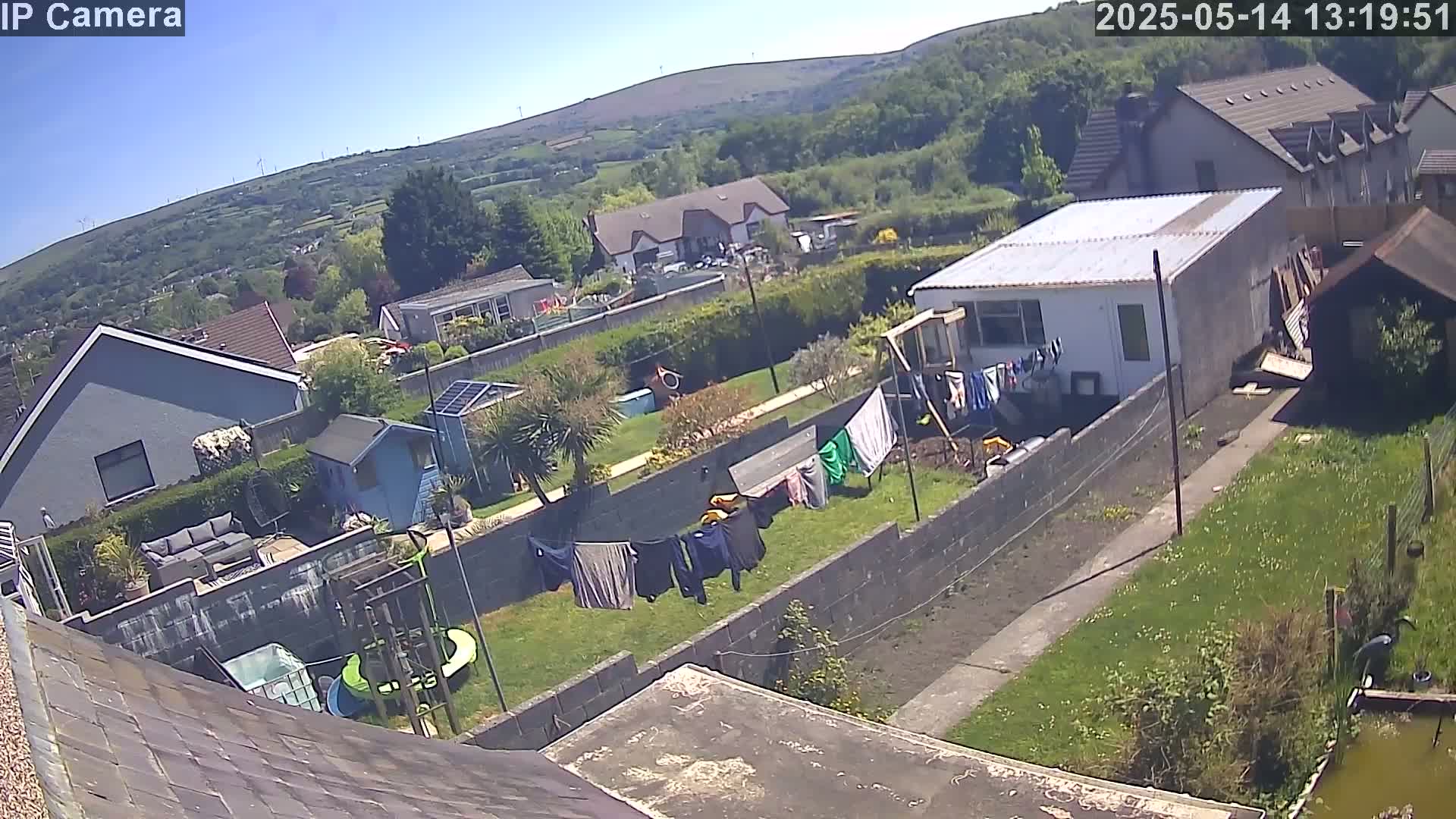 A sunny day view of a residential backyard with houses and green hills in the background, showing laundry drying on a clothesline and a small pond.